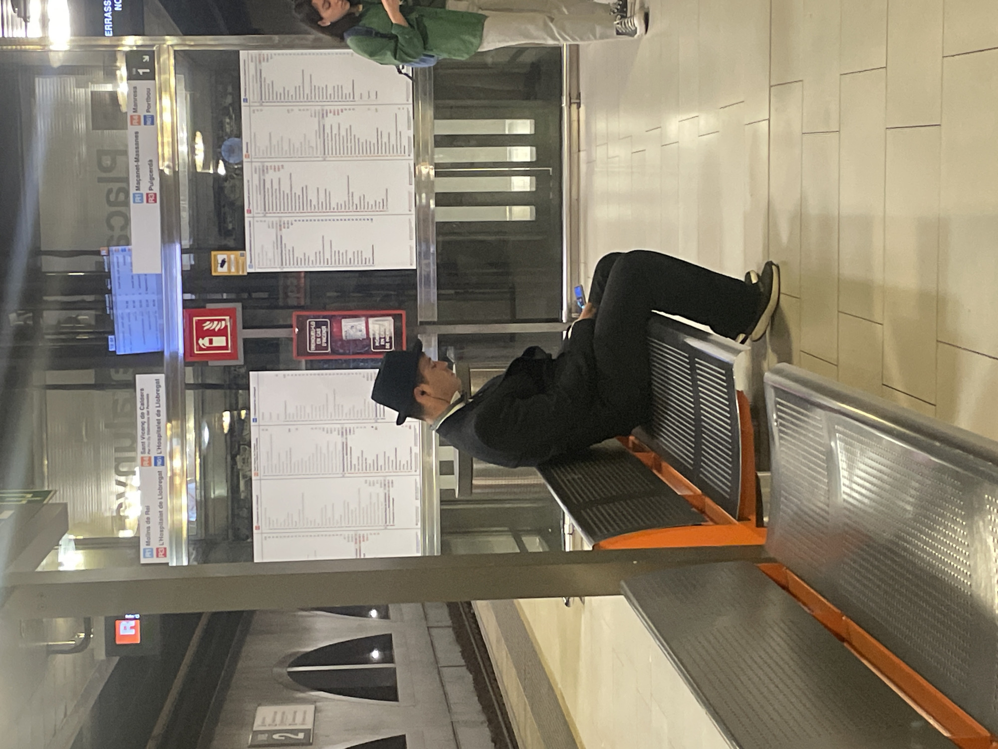 The image shows the interior of a Barcelona Metro station, Spain. A man in a black suit and hat is reclining on an orange-framed metal bench, looking at his cell phone. A woman in light pants and a green jacket is standing behind a train schedule board. There are train destination signs behind the board for destinations such as Sant Vicenç de Calders, Molins de Rei, L'Hospitalet de Llobregat, Manresa, Puigcerdà, and Portbou. A sign reads "Plaça".