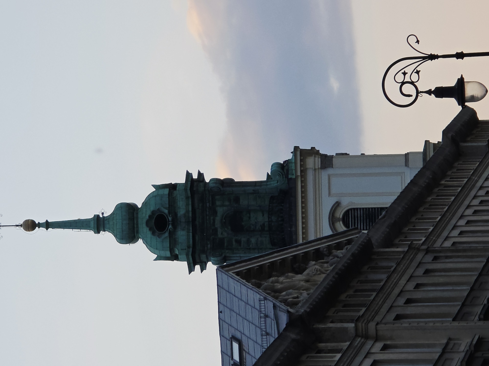 The image captures an upward-angled view of a building in Warsaw, Poland. A green spire-topped tower dominates the left side of the frame. To the right of the tower is a section of a white building with a rounded vent. At the bottom right corner, a portion of a building's roofline is visible. A wrought-iron lamppost is positioned at the upper right corner. The sky is filled with soft, muted colors, with clouds creating an ethereal backdrop. The image is rotated to the left.