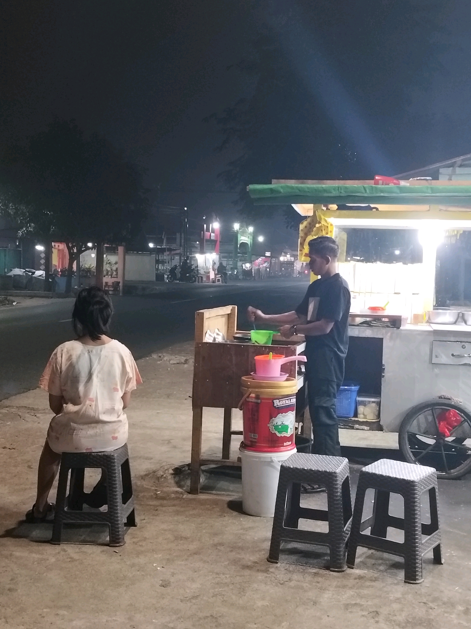A young Indonesian man, wearing a black t-shirt and black pants, stands behind a food cart in Arang Limbung, Indonesia, at night. He is preparing food, holding a ladle and a green bowl. In front of the cart are stacked plastic containers and a large red bucket. To the left of the food cart, a young Indonesian woman, wearing a light-colored patterned shirt, sits on a black plastic stool, facing away from the camera. Two other black plastic stools are empty in front of the food cart. The street in the background is illuminated by the lights of the food cart and distant buildings and streetlights. Motorcycles are parked along the street, and some people can be seen in the background. The scene suggests a typical street food vendor operation in a residential or commercial area at night.