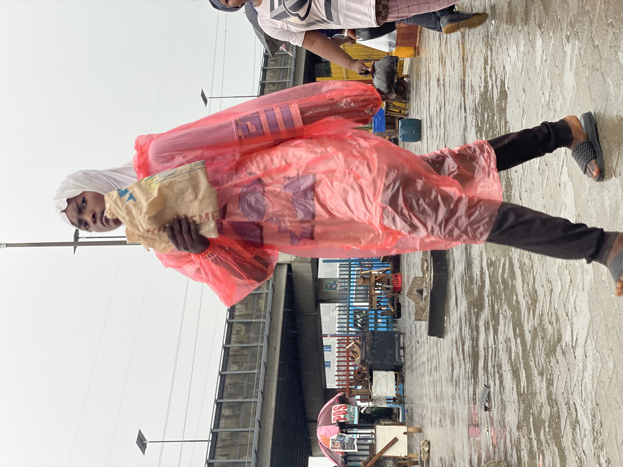 A young person wearing a white head covering and a transparent, orange rain poncho is seen holding a light-colored bag. They are wearing black leggings and open-toed sandals. The scene appears to be in Lagos, Nigeria, as suggested by the environment. The ground is wet, indicating recent rain, and there is an overpass in the background. Additionally, there are various objects, likely wares for sale, along the walkway. Another person is partially visible in the upper part of the frame.