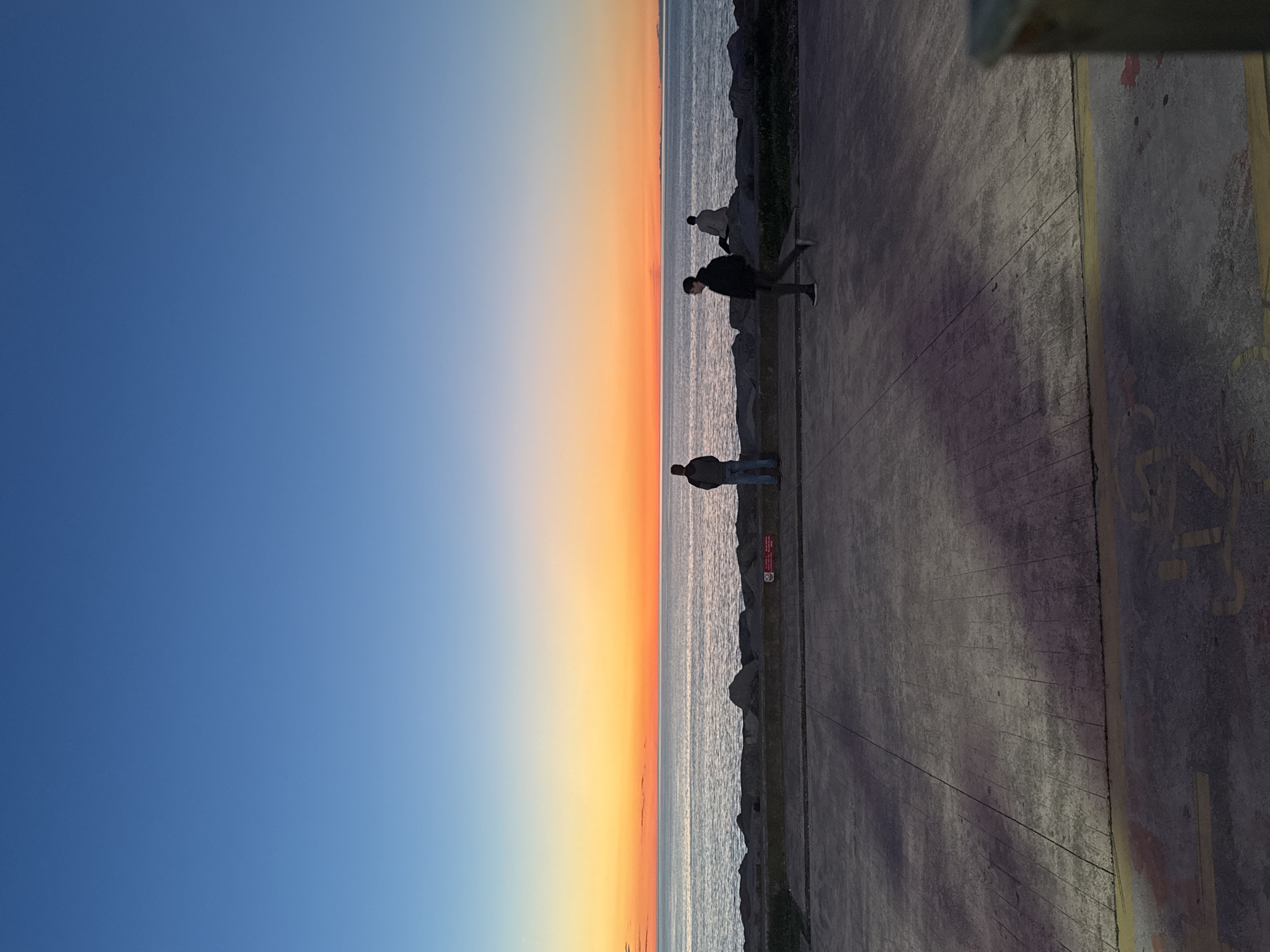 The image depicts a seaside scene at sunset in Batumi, Georgia. The sky transitions from blue to orange near the horizon, where it meets the sea. Three people are visible along a paved walkway bordering the water. The walkway appears to be constructed of concrete. A concrete structure exists in the upper right corner of the image.