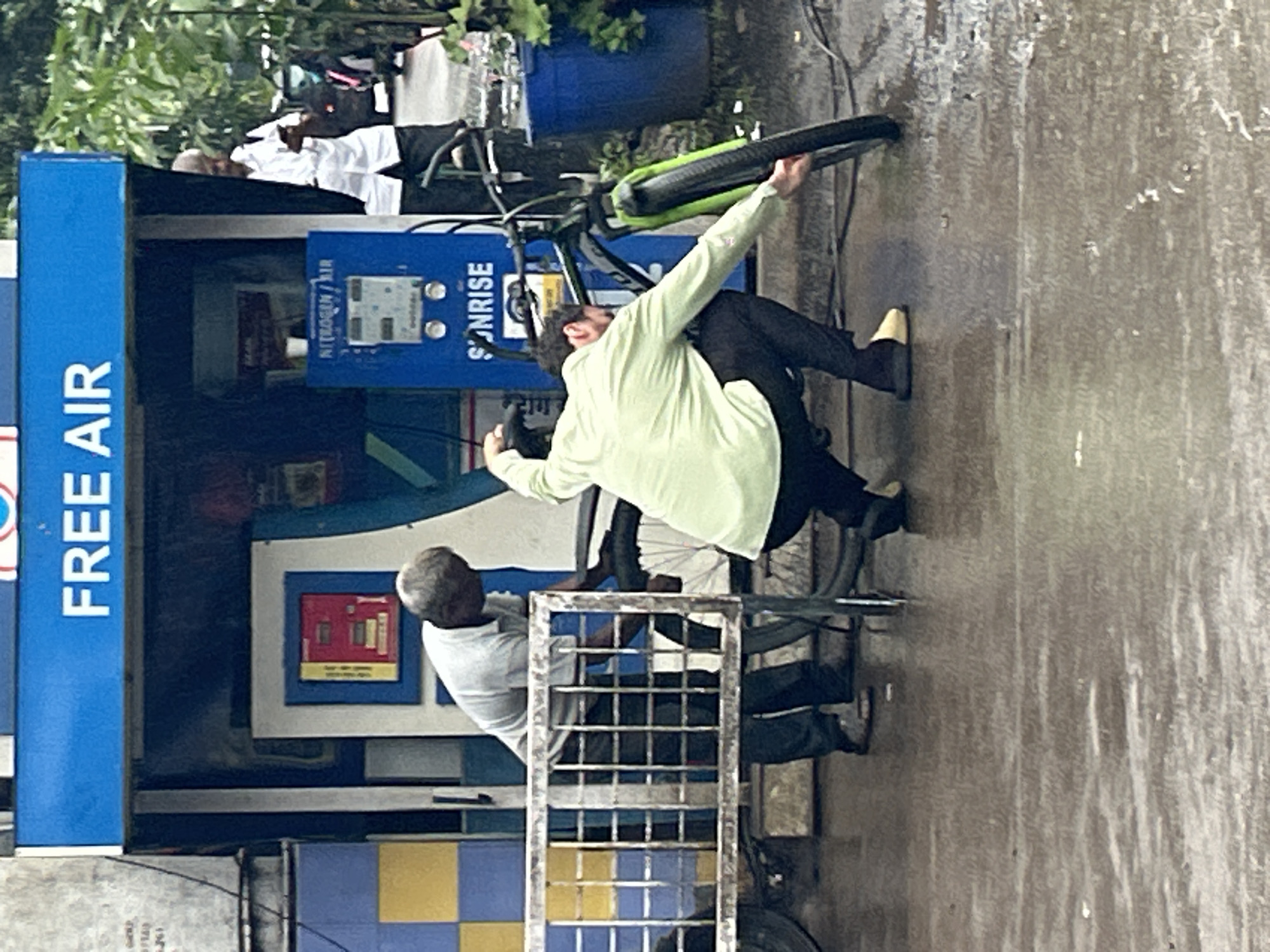 The image captures a street scene in Pune, India, likely at a roadside service station. The prominent blue awning displays "FREE AIR" in white lettering, indicating a service for inflating vehicle tires. In the foreground, a man wearing a light green shirt and black pants is bent over, attending to the front tire of a bicycle. He appears to be using a hose connected to a blue nitrogen/air machine visible behind him. To his left, another man in a light grey shirt and dark pants is also present, possibly assisting or observing. The ground is wet, suggesting recent rain, and a metal barrier with a grid pattern is situated between the two men. A blue barrel is visible to the right of the frame, and lush green foliage hints at the surrounding environment. The overall atmosphere is that of a functional, utilitarian service point in a bustling urban setting.