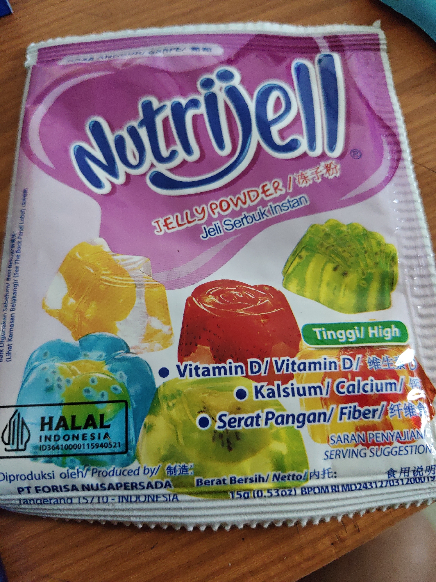 The image shows a close-up, top-down view of a package of Nutrijell jelly powder. The package is predominantly pink and purple with blue lettering. The brand name "Nutrijell" is prominently displayed in large blue letters. Below the brand name, it reads "JELLY POWDER / 冻子粉". Further down, it says "Jeli Serbuk Instan".
The front of the package features images of several colorful jelly molds: one yellow, one red, and two blue ones, one of which has green jelly with kiwi slices inside. A green jelly mold with kiwi is also visible on the right side.
In the bottom left corner, there is a rectangular black and white "HALAL INDONESIA" certification mark with the ID number "ID36410000115940521". Below this, text indicates "Diproduksi oleh/ Produced by/ 制造: PT FORISA NUSAPERSADA tangerang 15710 - INDONESIA".
Various nutritional and serving information is also present. A green banner reads "Tinggi/ High". Bullet points indicate "Vitamin D/ Vitamin D/ 维生素D", "Kalsium/ Calcium/", and "Serat Pangan/ Fiber/ 纤维". Text further down reads "SARAN PENYAJIAN/ SERVING SUGGESTION". Towards the very bottom, it states "Berat Bersih/ Netto/ 内托: 食用说明 15g (0.53oz) BPOM RI MD243127031200019".
The package is placed on a wooden surface, likely a table, which is visible in the background and slightly out of focus. The lighting suggests it is indoors. There are no people or actions depicted in the image, other than the presence of the product itself. The context of North Jakarta, Indonesia, is indicated by the text on the packaging.
