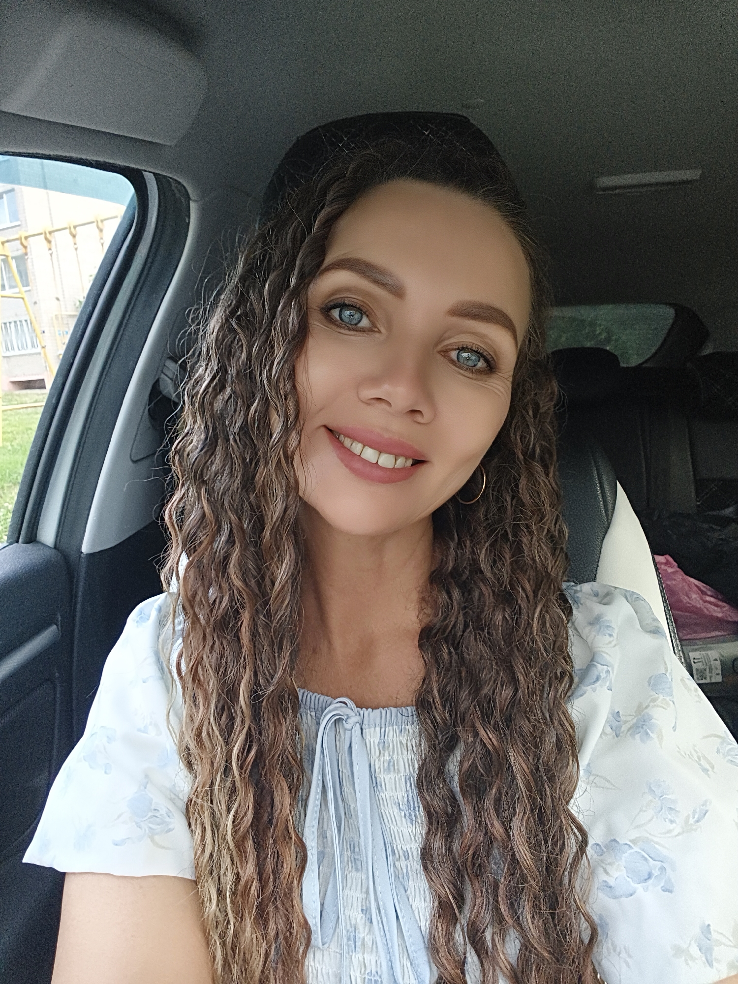A woman with long, curly brown hair and blue eyes smiles at the camera from inside a car. She is wearing a white top with light blue floral patterns. Her hair falls over her shoulders and down her front. The interior of the car is visible behind her, with a black seat and a glimpse of the outside through the window. Outside the car, a building with yellow scaffolding and a green lawn can be seen. The image appears to be taken in the city of Chaikovsky, Russia.