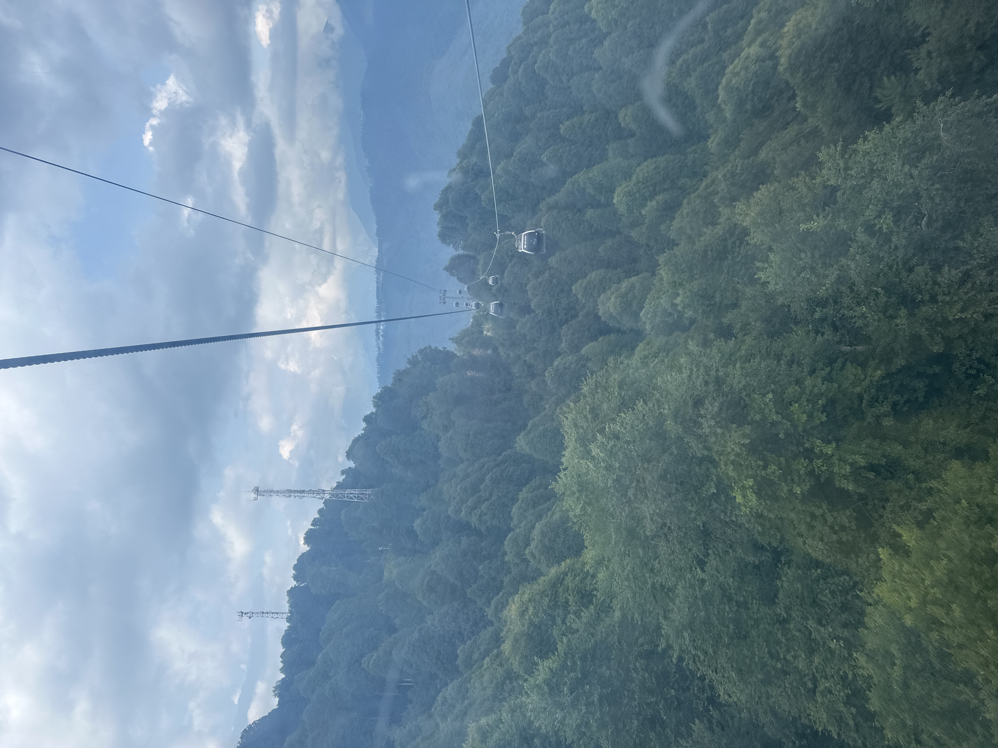 The image captures a view from a ski lift in Estosadok, Russia, overlooking a lush, forested mountainside under a cloudy sky. Multiple ski lift cabins are visible suspended from cables traversing the frame, ascending the steep incline. The mountains are densely covered with dark green trees, with lighter green foliage appearing in the foreground. Two tall, lattice-like communication towers stand on a ridge in the upper left portion of the image, further highlighting the mountainous terrain. The sky is a mix of light blue and grey clouds, suggesting overcast or potentially changing weather conditions. The overall scene conveys a sense of elevation and natural beauty within a mountainous resort area.