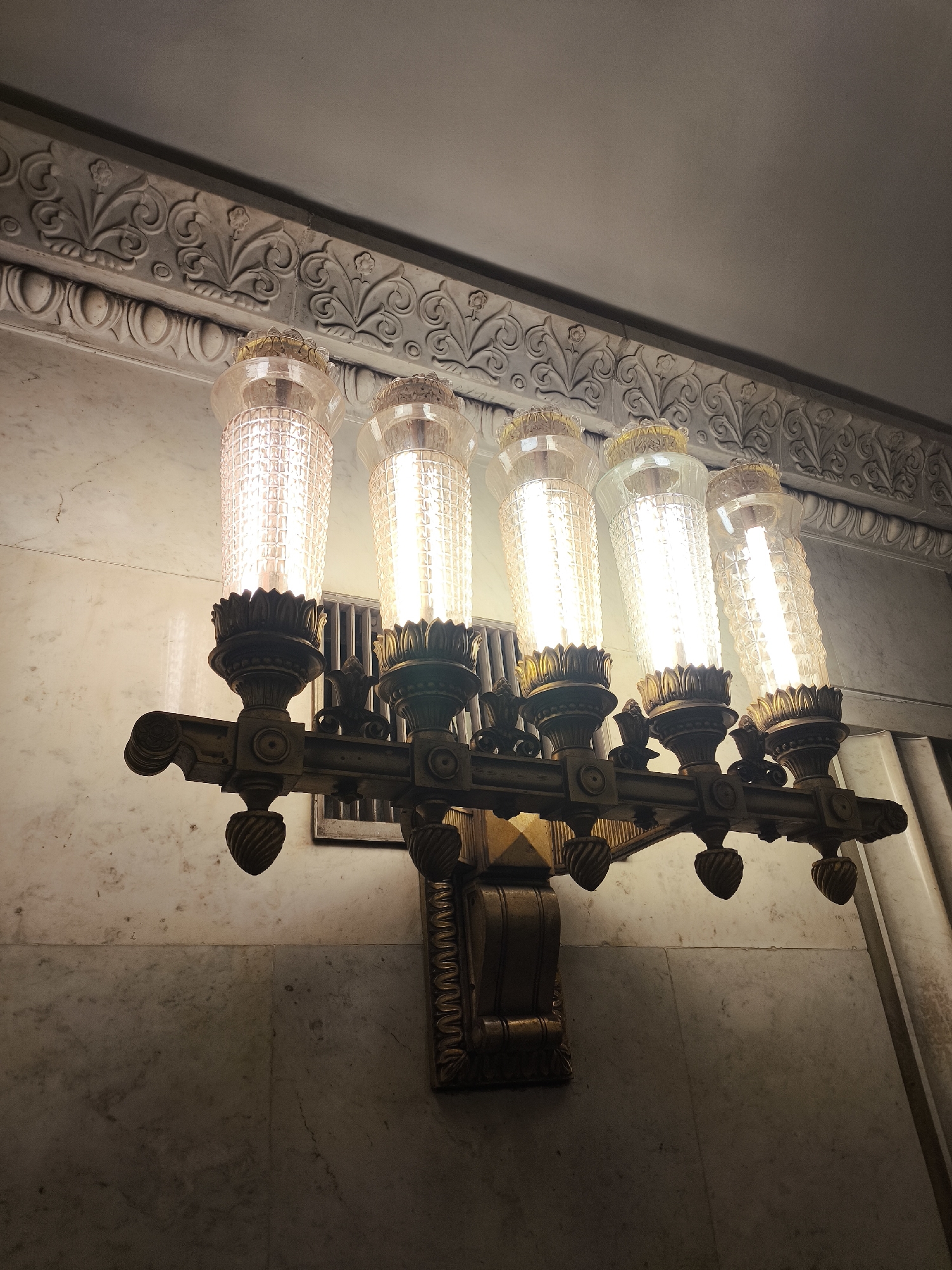 A low-angle, medium shot depicts a wall sconce with five light fixtures. The sconce is mounted on a wall made of marble. Above the sconce, there is an elaborate, decorative molding. The sconce is made of gold-colored metal and has a geometric, fluted design. Each light fixture has a clear glass cylinder with a diamond-shaped pattern. The lights are lit, casting a bright glow.