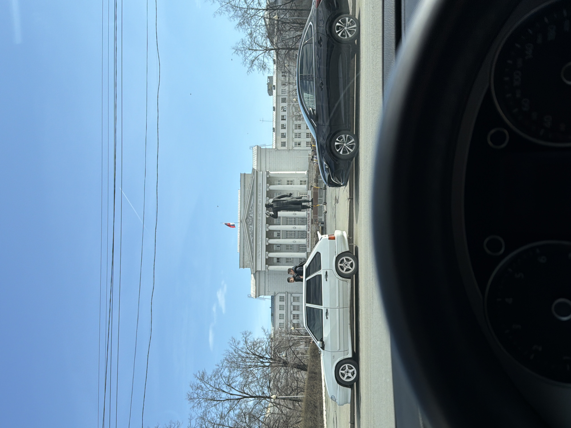 The image shows a street scene in Yekaterinburg, Russia, taken from inside a car, with the dashboard partially visible. A white Lada car is parked on the right side of the road, with two people visible inside. A black car is parked in front of the Lada. In the background is Ural Federal University, a white building with columns and a statue of Dmitry Mendeleev in front of it. A Russian flag is waving atop the building. Power lines run across the blue sky. The trees are bare.