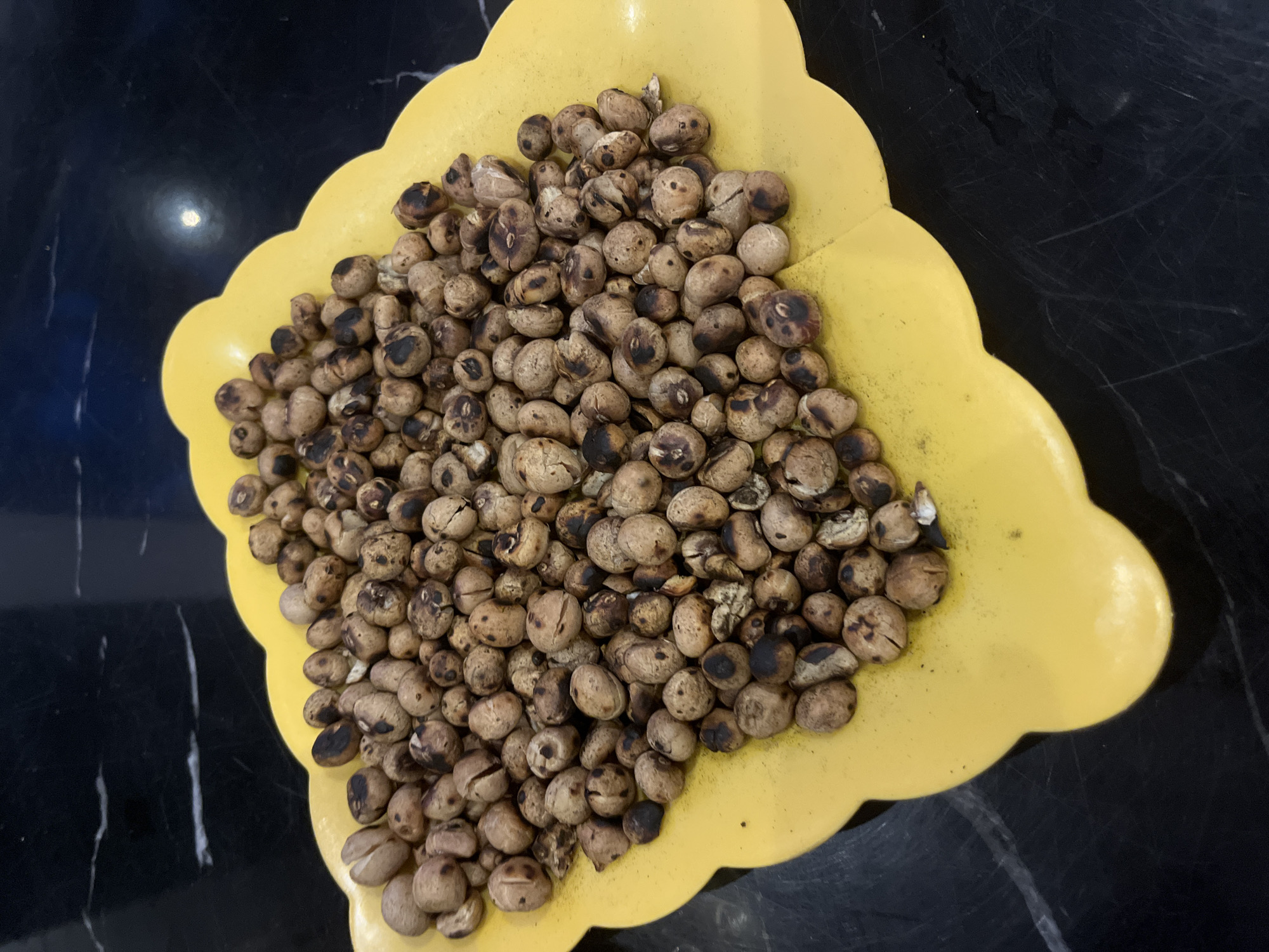 A yellow, decorative bowl filled with roasted tiger nuts sits on a black marble surface. The nuts are spherical and vary in shade from light brown to nearly black, appearing charred in spots. The surface is reflective, showing a glare of light.
