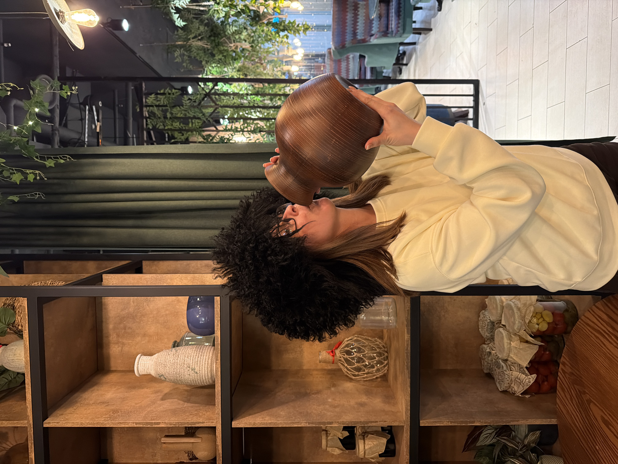 A woman wearing glasses and a furry black hat is laying down next to a wooden shelf with various items. She is holding a brown ceramic vase to her mouth as if she is drinking from it. The shelf contains items such as blue and white vases, a net-covered sphere, jars filled with food, and a small plant. Behind the woman and the shelf is a green curtain and a second-story balcony area with plants and lighting fixtures. The location appears to be inside a building. The city and specific location are unknown.