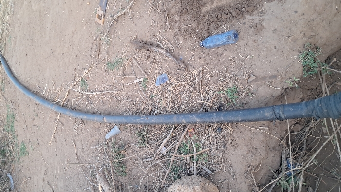 The scene in Walu, Kenya, depicts a dry, reddish-brown earthen ground with sparse green vegetation and scattered debris. A long, black, flexible pipe, approximately 7-10 cm in diameter, extends diagonally across the frame from the upper right toward the lower left. The pipe appears to be partially laid within a shallow trench or furrow in the soil, particularly noticeable on the right side and near the bottom where it transitions into a corrugated section.
Numerous objects are dispersed around the pipe. These include:
* Several dry twigs and branches, some forming small piles.
* Two translucent plastic bottles, one clear and one blue, are visible on the ground. The blue bottle near the lower right appears to be partially crushed or cut.
* A section of the black pipe, approximately one-third of the way from the bottom, is wrapped with black adhesive material or tape.
* A large, light-colored rock is positioned on the far left of the frame.
* A hand saw with a light-colored wooden handle and a dark metal blade is lying on the ground near the top right corner.
No individuals or active interactions are visible. The environment suggests an outdoor, possibly arid or semi-arid, area with evidence of recent activity such as digging or material transport.