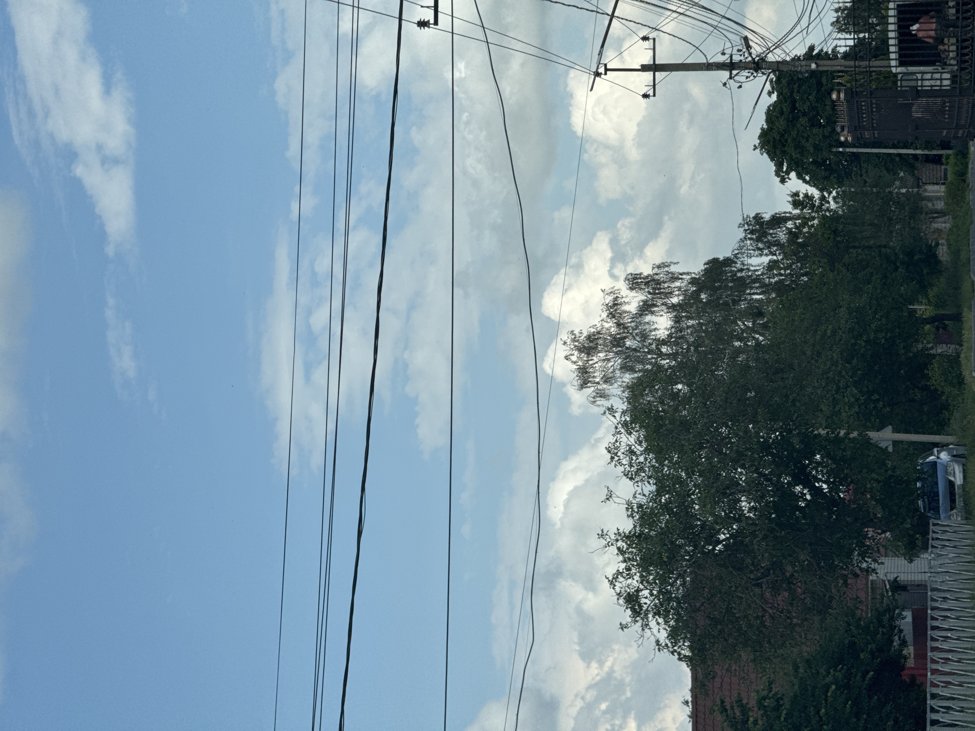 The image depicts a street scene with overhead power lines dominating the upper portion of the frame. Below the power lines, a bright blue sky is visible, interspersed with fluffy white clouds. Trees, some appearing to be birch with drooping branches, line the street. A building with a red-tiled roof is partially visible behind the foliage on the left. In the foreground, a white picket fence runs along the side of the street. A silver car is parked near the fence. On the right side of the image, a utility pole with numerous wires is present, alongside a decorative black metal gate. A person can be seen through the gate, appearing to be engaged in activity within a gated area. The scene is set in Mikhaylovsk, Russia.