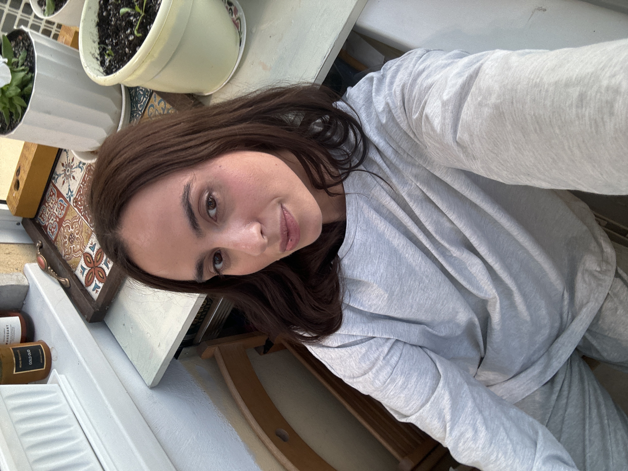 A woman with long brown hair, wearing a light gray sweatshirt, is taking a selfie. In the background, there are potted plants and a decorative tile tray on a white surface, likely a windowsill or shelf. A wooden chair is visible in the lower part of the frame. The location cannot be determined to be Krasnoyarsk, Russia based on the visual content of the image.