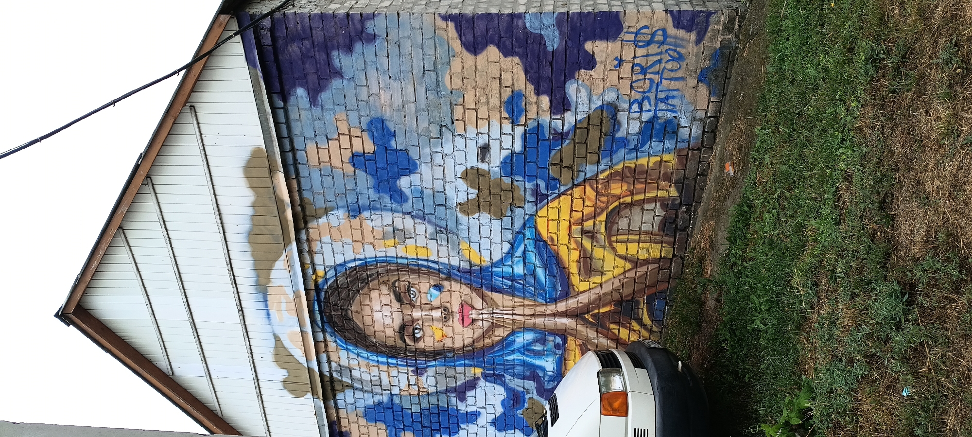 The image shows a mural painted on the brick wall of a building, likely a garage or storage structure. The mural depicts a young woman with her hands clasped in prayer, wearing a blue headscarf and a yellow garment. Her eyes are wide and looking directly at the viewer. Abstract blue and brown shapes form the background, which is painted over the brickwork. To the right of the mural, the text "BORIS TATTOO" is visible in blue spray paint. In the foreground, the front of a white van is partially visible on the left, and overgrown grass covers the ground. The building's white-paneled gable is visible at the top of the image. The scene appears to be located outdoors in an urban or semi-urban environment.