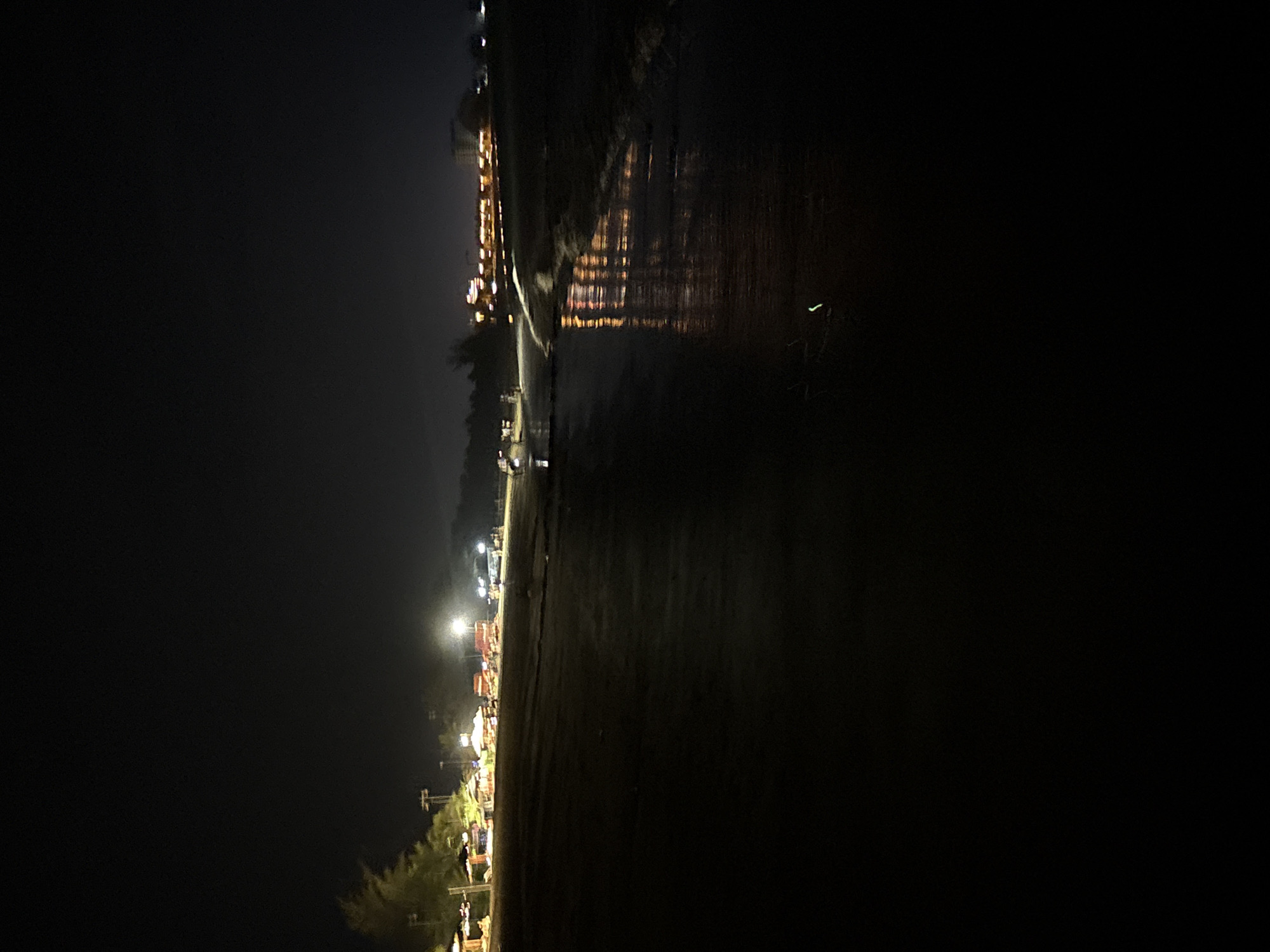 The image displays a horizontal nighttime scene along a waterfront, likely a beach or riverbank, in Dong Hai Ward, Vietnam. On the left side, a strip of land is brightly illuminated by numerous artificial lights. These lights appear to originate from buildings, market stalls, or streetlights, creating a vibrant glow that suggests a bustling promenade or commercial area. Indistinct figures, likely people, can be seen within this illuminated zone, hinting at human activity.
Separating this lively land strip from the main body of water is a dark, unlit shoreline. To the right, a large expanse of water dominates the scene, reflecting the artificial lights from the left with striking clarity. These reflections form shimmering, elongated streaks of orange, yellow, and white across the dark water's surface. A distinct, bright green light or object is visible floating or positioned within the water towards the far right. The sky above is uniformly dark, indicating a clear night. The overall impression is one of a calm yet active evening, possibly an outdoor market or leisure area beside the water. No visible text is present.