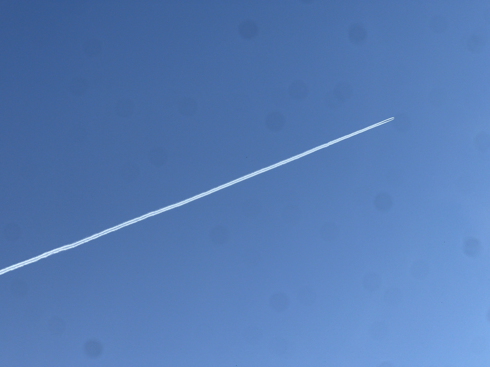 The image shows a contrail in a clear, blue sky. No buildings or ground features are visible, so it is not possible to determine if the image was taken in Budva, Montenegro, or any other specific location. There are no people, subjects, objects, actions, or interactions discernible beyond the contrail itself. The environment consists solely of the blue sky.