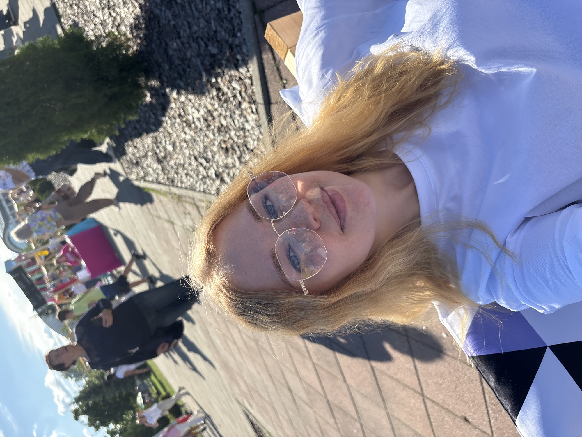A woman with blonde hair and glasses is sitting on a bench. She is wearing a white t-shirt. The bench has a checkered pattern in black, white, and purple. In the background, people are walking around in an outdoor area with trees and stalls. The location appears to be the city of Rusanovo, Russia, likely during a festival or event, given the stalls and the number of people present. The sun is shining, casting shadows on the ground.