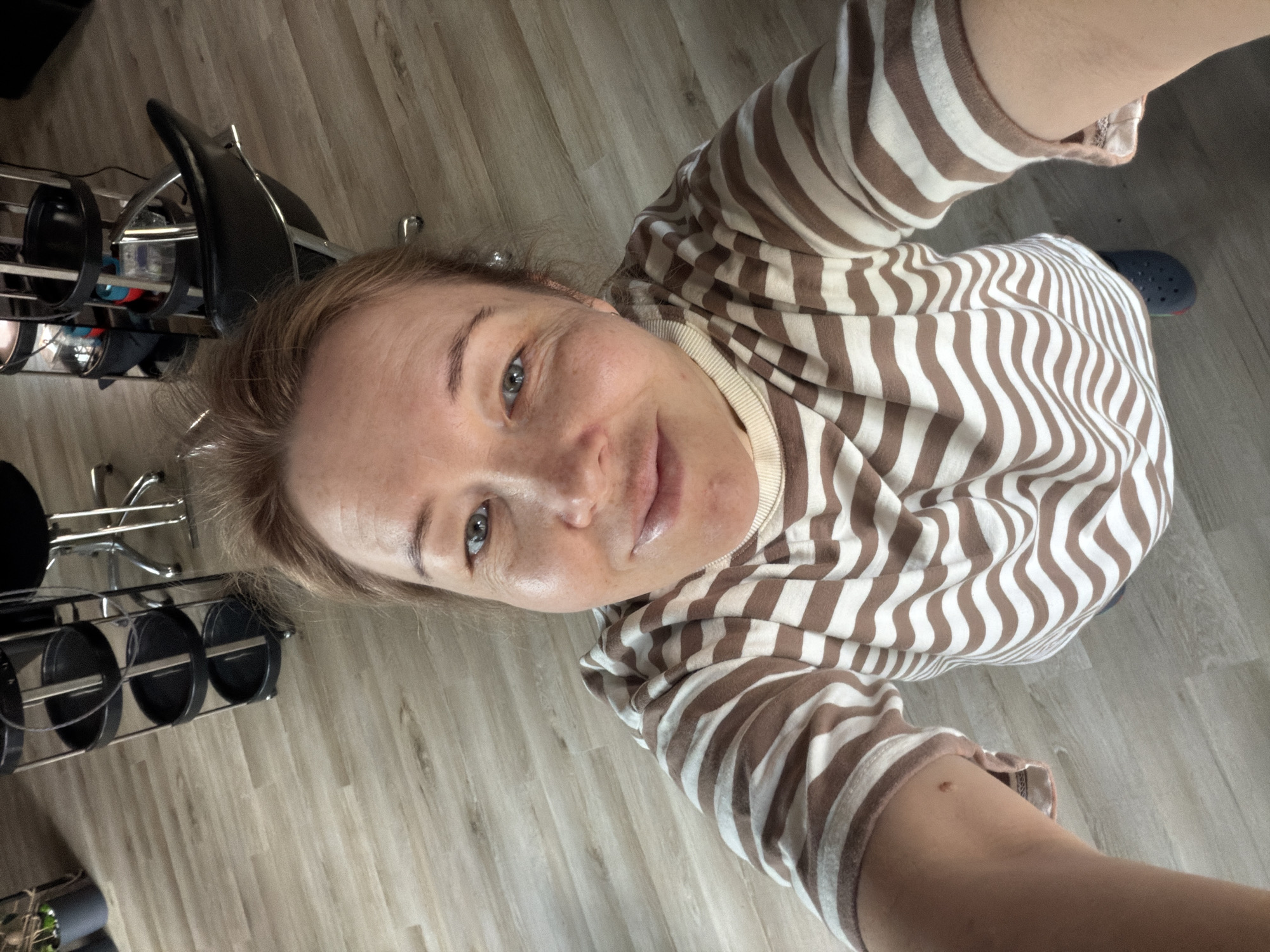 A woman with fair skin and light brown hair is taking a selfie from a high angle. She is wearing a brown and white striped long-sleeved shirt. A portion of a blue Croc shoe is visible near her. Behind her, there is a barber's station with salon chairs and styling tools. The floor is light-colored wood-grain laminate. It is not possible to confirm the location as the city of Pervomaysky, Russia from the image.