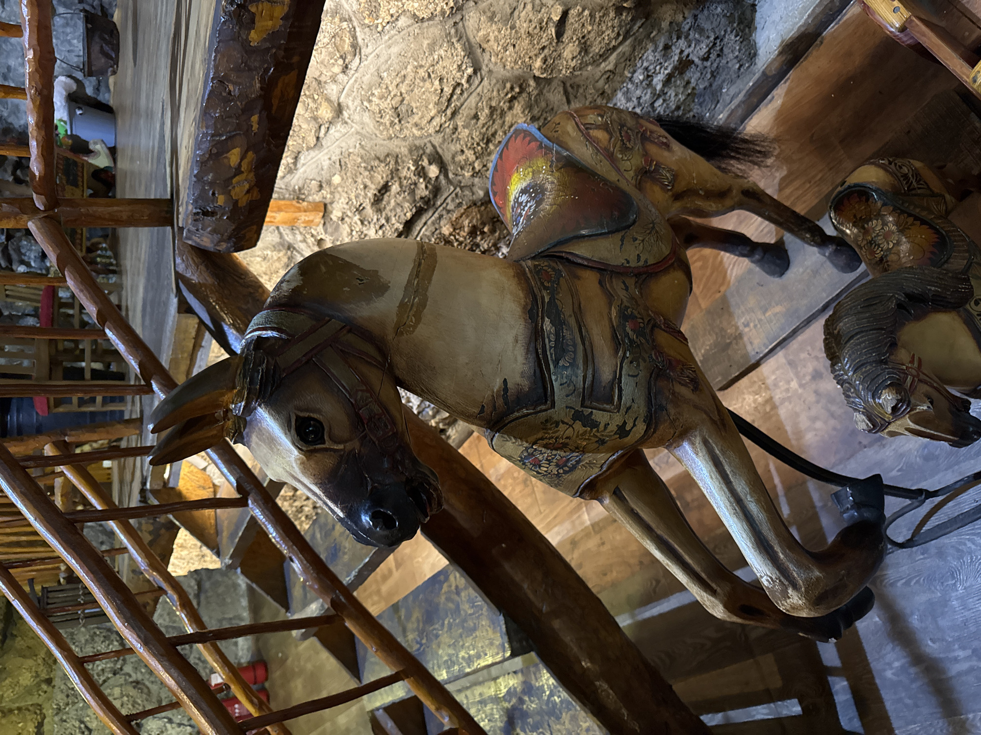 The image shows two vintage carousel horses inside a building, likely in Podgorica, Montenegro. One horse is positioned closer to the viewer, its head prominent, with details of its bridle and painted body visible. A wooden staircase is on the left side of the frame, leading upwards, and a stone wall is visible in the background. The interior suggests a rustic or historical setting. The presence of the carousel horses suggests the location might be a museum or a building with decorative elements from a historical amusement park.