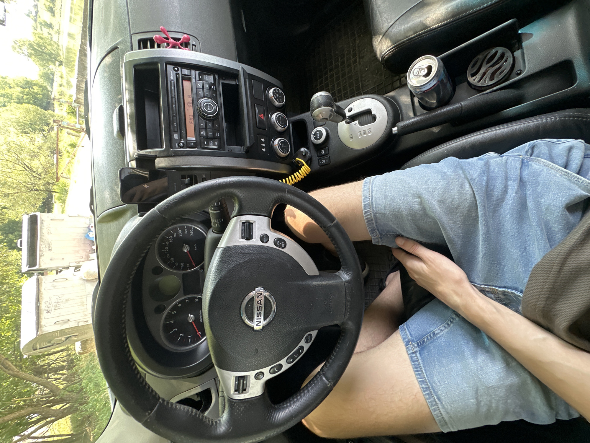 The image shows the interior of a Nissan car, viewed from the driver's perspective. The dashboard includes a steering wheel with Nissan branding, a radio, climate controls, and a gear shifter. A person's legs, wearing denim shorts, are visible in the foreground, with one arm resting on their lap. A can of soda is placed in the center console. Outside the car, through the windshield, two large dumpsters and trees are visible, suggesting an outdoor, possibly semi-rural or industrial setting. The image is likely taken in Makiv, Ukraine, based on the provided context.