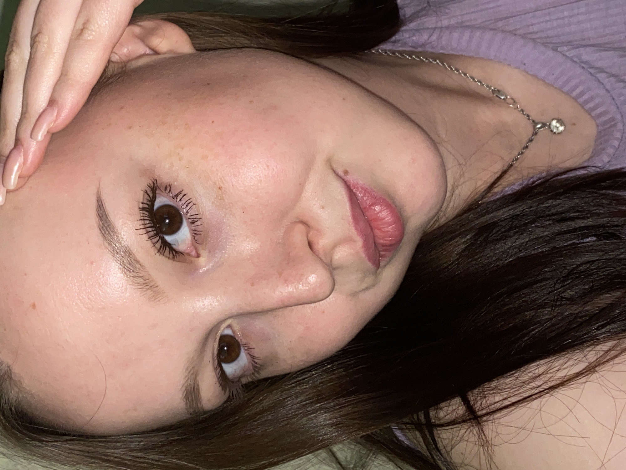 A close-up, tilted portrait features a young woman's face, captured indoors in what appears to be a relaxed setting in Chaikovsky, Russia. She is positioned on her side, looking towards the viewer with a soft, subtle expression, possibly a gentle smile. Her fair skin shows a scattering of light freckles. She has brown eyes, framed by long, dark, mascara-enhanced eyelashes and well-defined dark eyebrows. Her lips are a soft pink.
One of her hands, with manicured nails painted in a light, neutral shade, rests near her forehead and ear. Her dark brown hair is visible around her head. She is wearing a light purple ribbed top, and a delicate silver chain necklace with a small, round clear gem pendant is visible around her neck, resting on her shoulder. The background is softly blurred, showing the purple fabric of her top and darker areas, suggesting an indoor environment like a bed or couch. The lighting is soft and even, highlighting her features. No text is visible in the image.