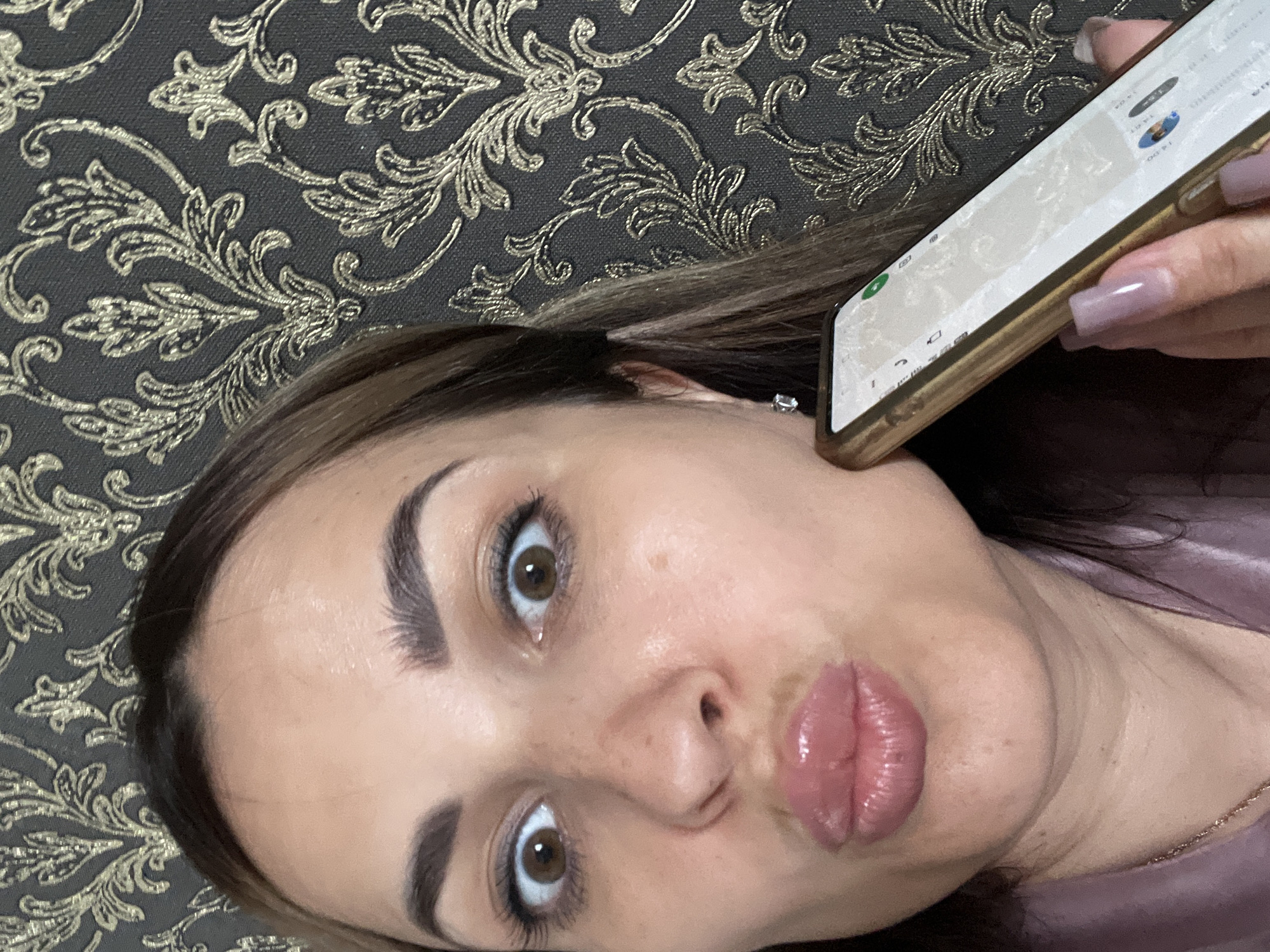 A selfie shows a woman with dark brown hair and brown eyes, pursing her lips. She is holding a gold-colored smartphone in her right hand, which is positioned near her cheek. The woman is wearing a light purple satin robe and a small diamond earring. The background is a dark gray textured wallpaper with a gold damask pattern. The scene is indoors and appears to be in a domestic setting.