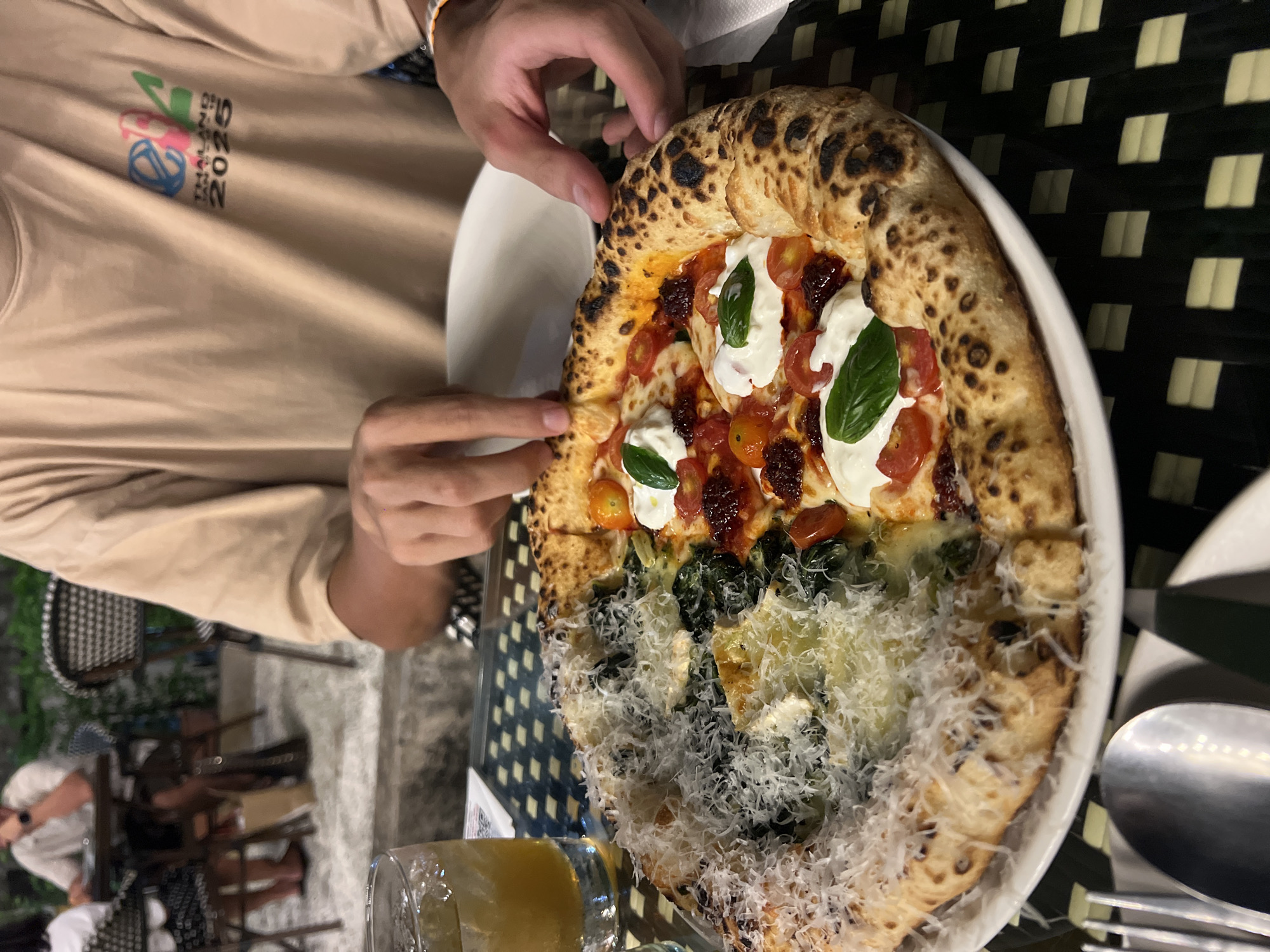 The image shows a person holding a pizza on a white plate. The pizza is half covered with fresh tomatoes, cheese and basil, while the other half is topped with spinach and grated cheese. The pizza is positioned in the center of the frame. The person's hand is visible, holding a slice of the pizza. The person is wearing a beige t-shirt. The table beneath the plate has a patterned surface. A glass of amber-colored liquid sits near the bottom of the frame. Forks and Spoons are seen to the right of the frame. The environment appears to be an outdoor dining area. The city and specific location are unknown.