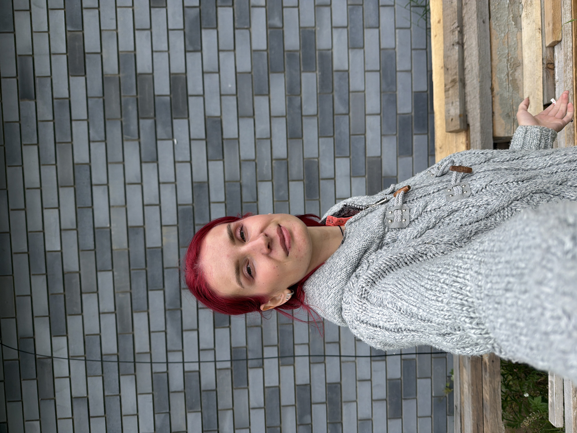 An outdoor portrait captures a person with vibrant red hair, looking directly at the camera with a gentle smile. They are wearing a thick, light grey cable-knit sweater, adorned with wooden toggles and grey leather or faux leather loops. A small silver earring is visible in the person's left ear. Their right hand is slightly raised and open, holding a small white cylindrical object, which appears to be a cigarette.
The background is divided, featuring a wall of rectangular grey tiles arranged in a staggered pattern on the left, and a rustic wooden structure, possibly a fence or bench, composed of vertical planks on the right. Green foliage is visible beneath the wooden structure.
The scene takes place during the daytime, under soft, diffused lighting, suggesting an overcast sky or shaded conditions. The heavy sweater indicates cool weather. The person's expression is calm and pleasant. There is no visible text in the image. The location context provided is Chaikovsky, Russia.