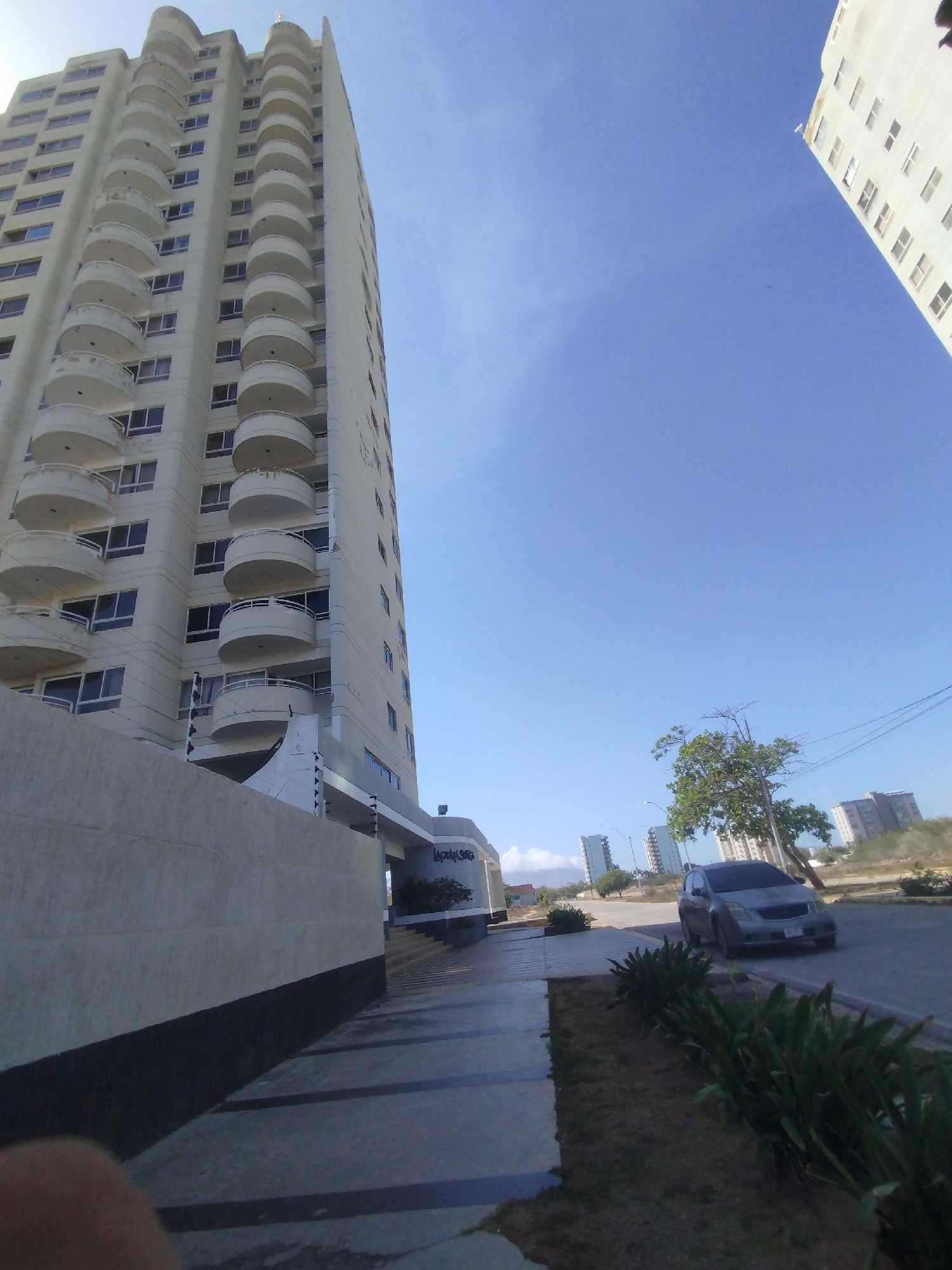 The scene depicts a street view in Porlamar, Venezuela. A tall, light-colored apartment building with rounded balconies dominates the left side of the frame. A lower wall with a dark base runs along the sidewalk in front of the building. The sidewalk is paved with grey tiles. A grey vehicle is parked on the street next to a grassy area with green plants. In the background, there are other buildings and trees. The sky is blue and mostly clear.