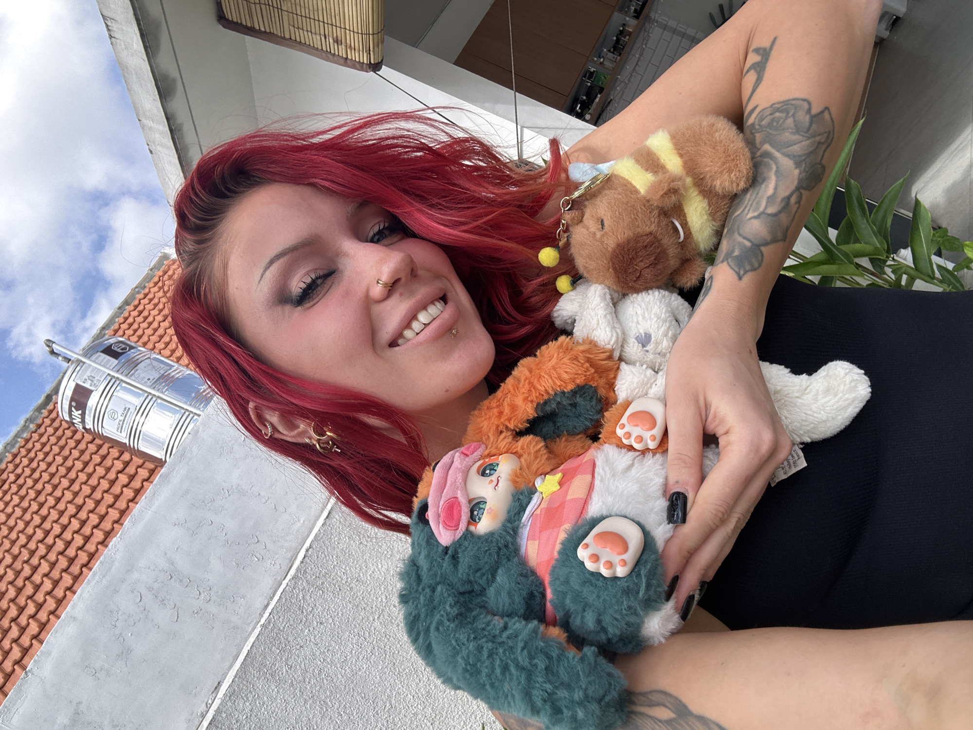 A woman with vibrant red hair and extensive arm tattoos is holding several plush toys. She is wearing a black tank top and has a nose ring and several ear piercings. One plush toy is predominantly teal with orange accents and appears to be a character with large eyes. Another is an orange and white creature, and a third is a small, brown, bee-themed plush toy attached to a keychain. The woman is smiling and looking at the camera.
The background suggests an outdoor setting, possibly a rooftop or balcony. Elements visible include a terracotta tiled roof, a grey textured wall, a large silver water tank, and bamboo blinds on a building. Green foliage is also present in the foreground. The overall impression is that of a casual, personal moment captured outdoors.
The location is identified as North Kuta, Indonesia, suggesting a tropical or subtropical climate indicated by the presence of lush greenery and open-air elements.