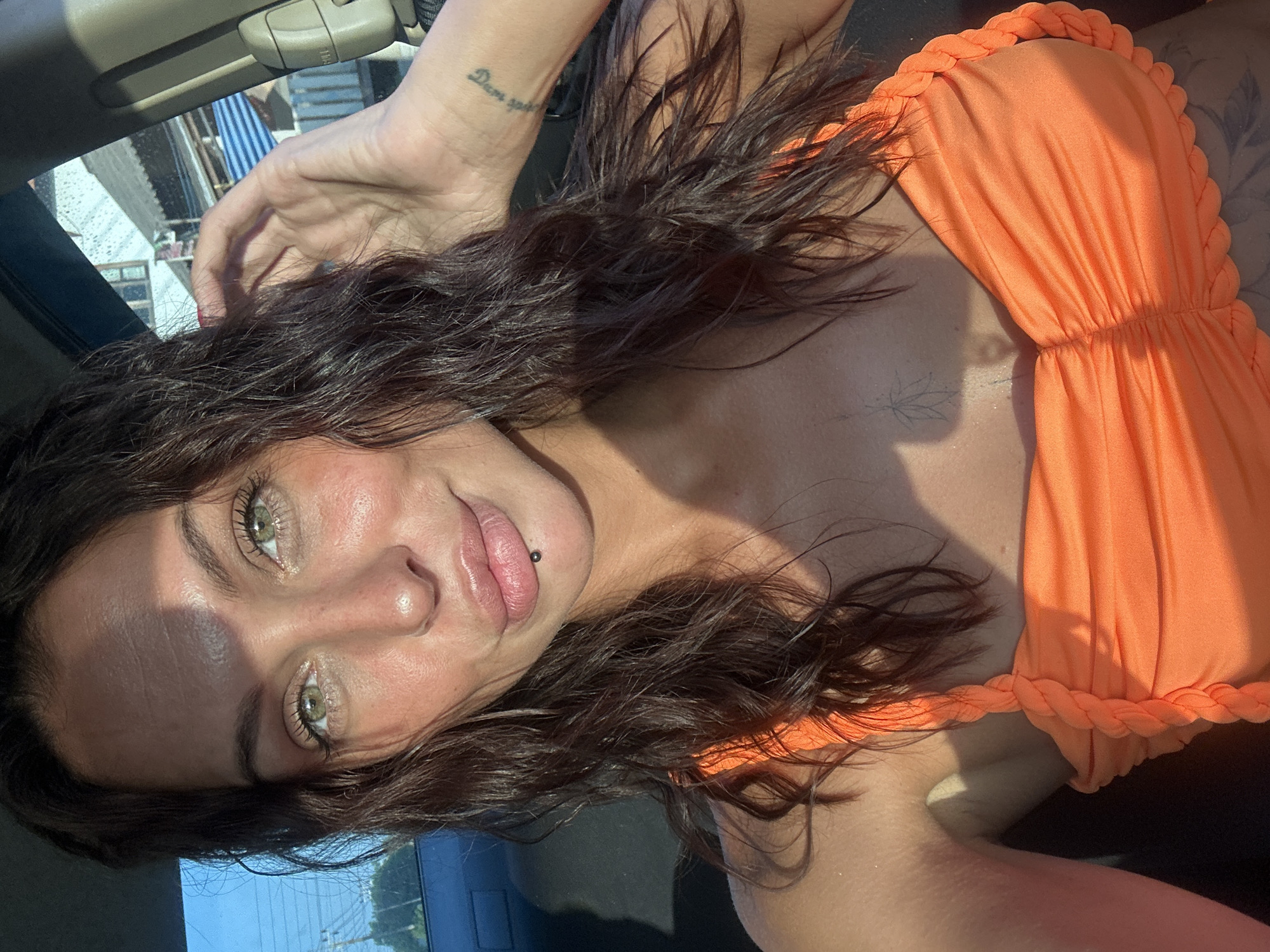 In a vehicle, a young woman with dark wavy hair and green eyes looks directly at the camera. She is wearing an orange ruched bikini top. Her left hand is raised, touching her hair. A sun shadow falls across her face and body. In the background, through the car window, houses with tiled roofs and blue and white striped awnings are visible, suggesting an urban or suburban setting in a warm climate.