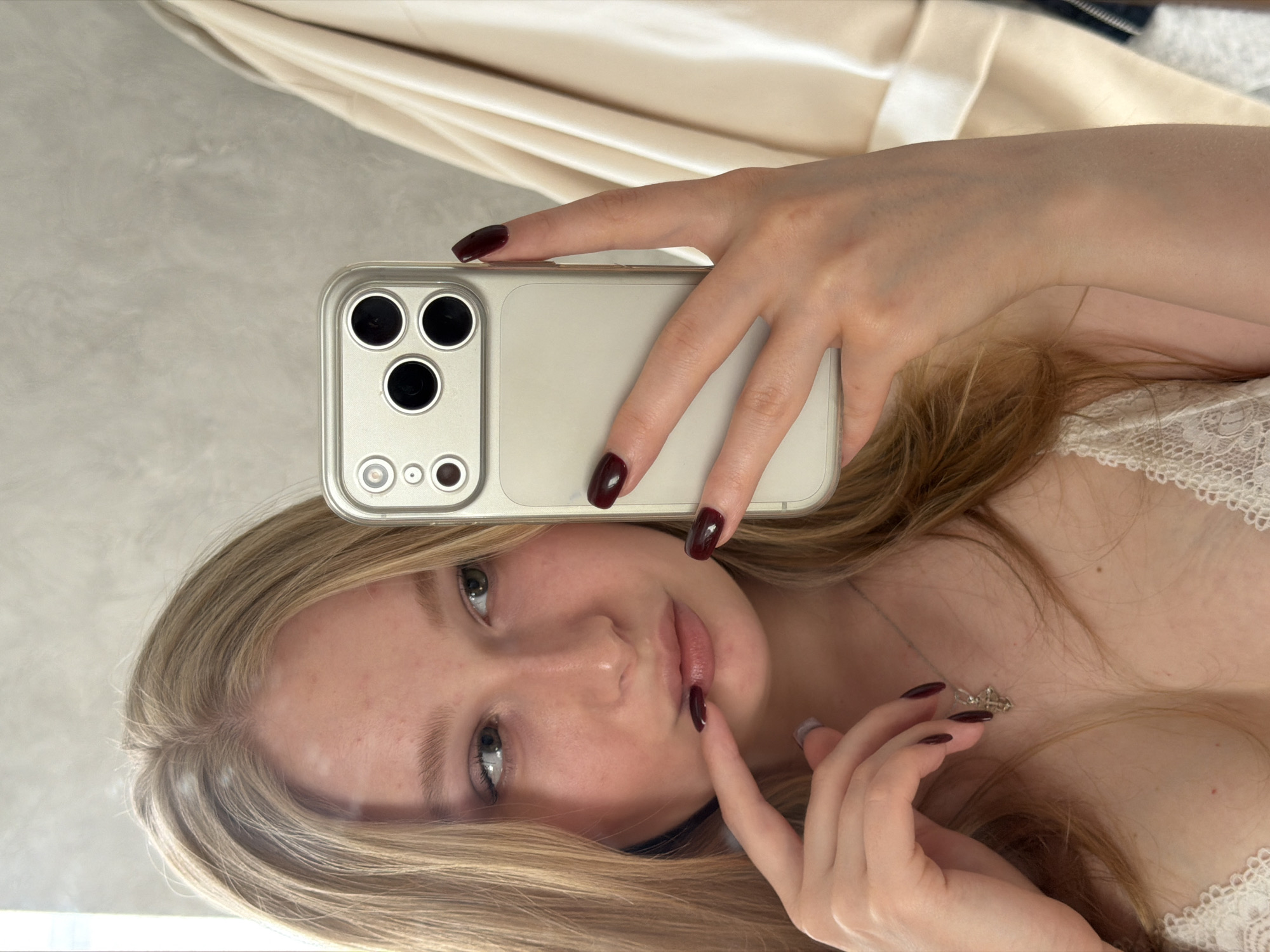 A woman is taking a selfie in front of a mirror. She is holding her phone with her left hand and her right index finger is on her lip. She has blonde hair and is wearing a white lace top. The location is not discernible.