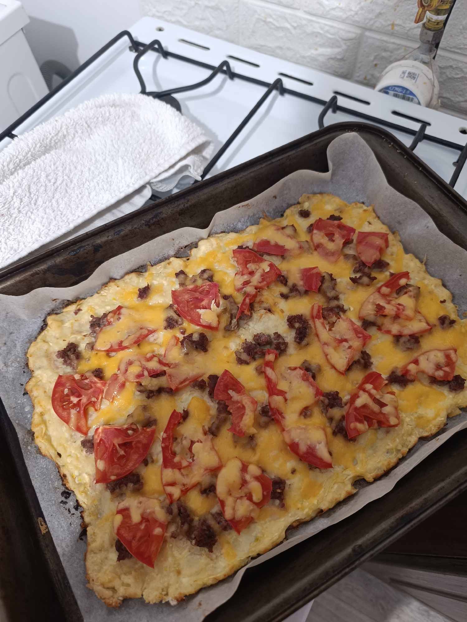 A close-up, high-angle shot shows a freshly baked keto pizza on a baking sheet lined with parchment paper. The pizza has a golden-brown crust, melted yellow cheese, crumbled ground meat, and slices of red tomato. The baking sheet is placed on a white stovetop with black grates. To the left of the stovetop is a white folded towel. In the background, a white brick-patterned wall is visible. A gas meter with a blue and white label is attached to the wall above the stovetop. This scene appears to be a kitchen setting, possibly in Taraz city administration, Kazakhstan, given the provided context.