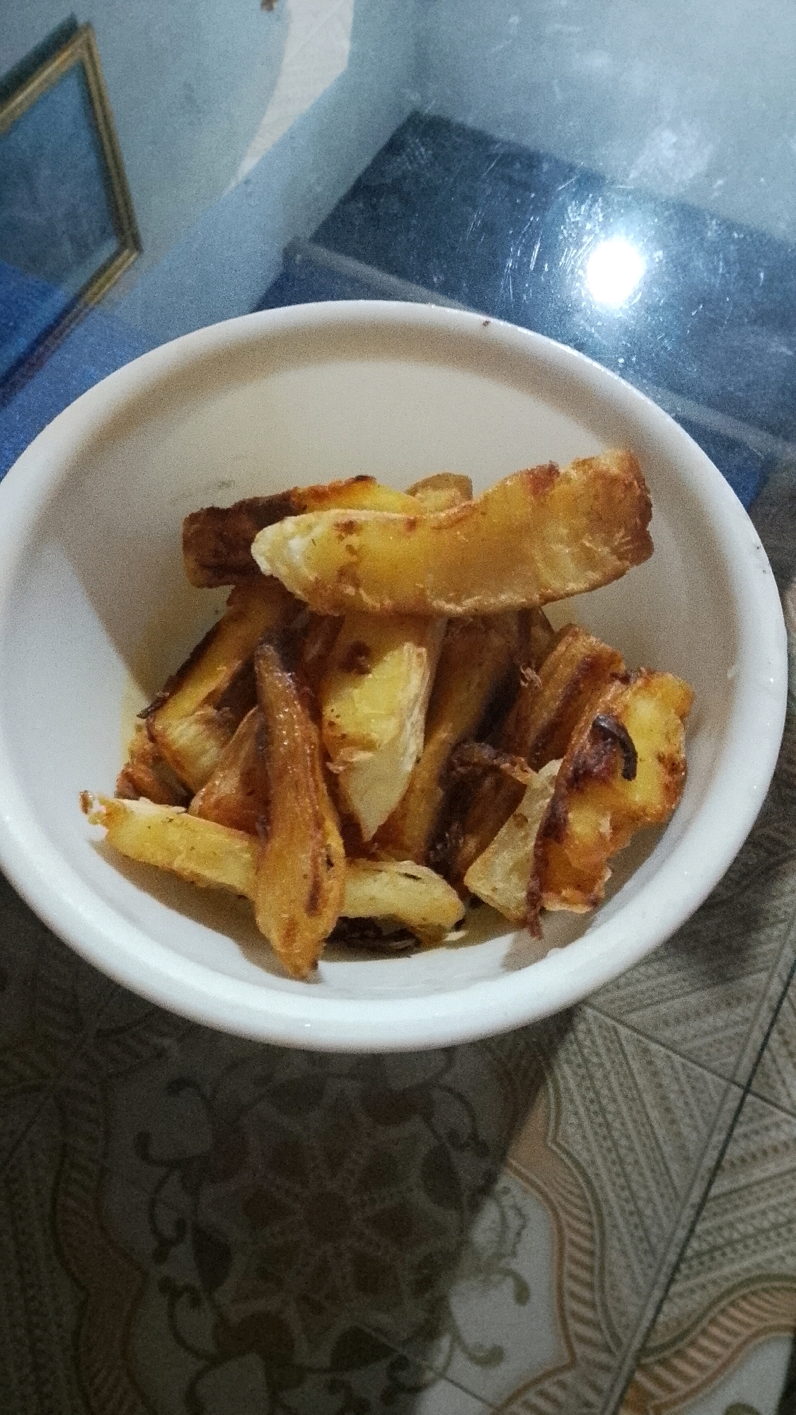 A white bowl filled with potato wedges sits on a patterned table. The potato wedges are golden brown and appear to be fried or roasted. The table surface is a light beige with a repeating pattern of dark brown and light tan swirls. In the background, a blue surface is visible, with a rectangular object with a golden frame. A bright light source reflects off a dark surface in the upper right corner. The overall lighting is dim, with a slight overhead glow. The setting is likely a home interior in Mulyo Haji, Indonesia, given the context provided.