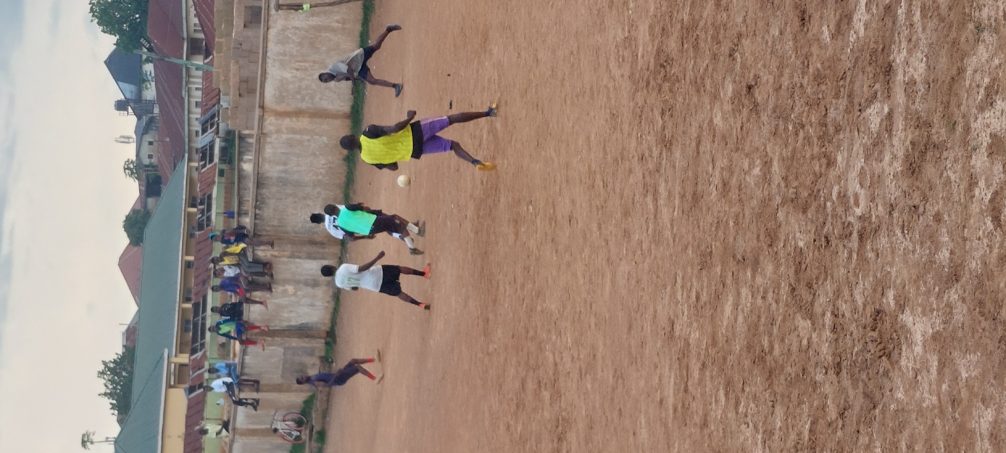 The image depicts an outdoor scene of a football match being played on a dirt field, possibly in Bukuru, Nigeria. Several young men are actively engaged in the game. One player in a bright yellow vest and purple shorts is dribbling the ball. Another player in a light blue vest and black shorts is nearby. Two players in white jerseys and dark shorts are also visible on the field. Further in the background, a player in a dark blue jersey is running. Several spectators are seated on a low concrete wall in the background, watching the game. The environment consists of a dusty, uneven dirt field with a concrete wall and a backdrop of buildings with corrugated iron roofs, indicative of an urban or semi-urban setting. The sky is overcast. A bicycle is leaning against the wall in the far left of the frame.