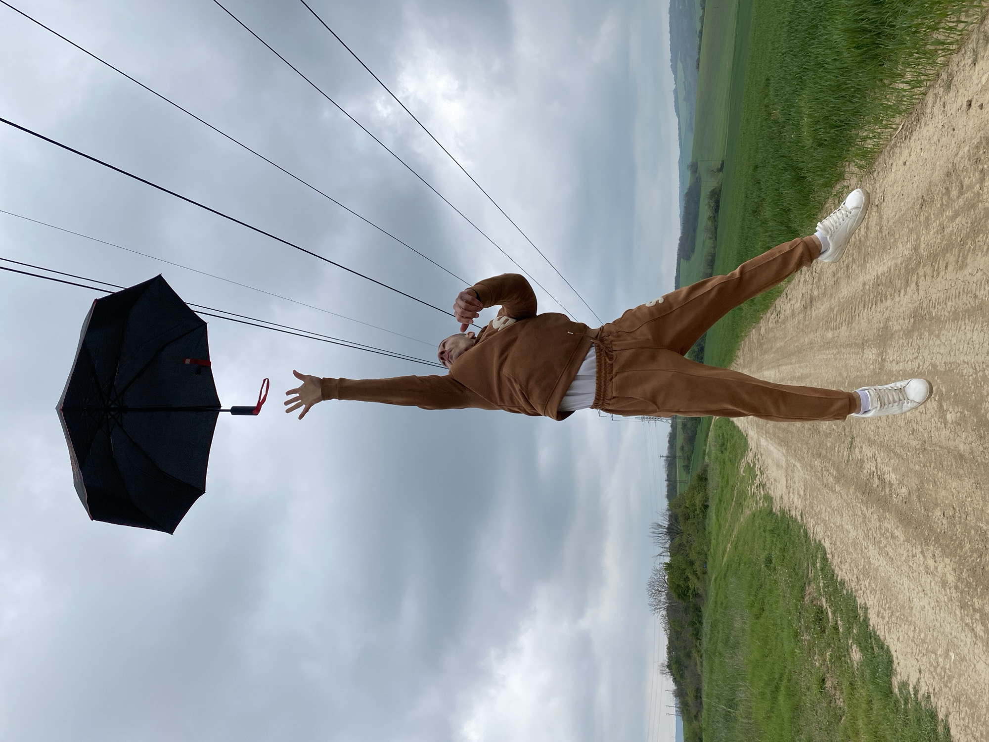 The image is captured with a horizontal perspective, showing a person positioned as if floating or suspended parallel to the ground. The individual wears a brown matching tracksuit with "SWEDEN" visible in white text on the left pant leg, along with white sneakers. Their right arm is fully extended towards a black umbrella with red detailing, which is positioned slightly below their hand, suggesting an interaction with it. Their left arm is bent, with the hand near their head. The person's expression is not clearly visible.
The backdrop features an overcast, grey sky filling the upper portion of the frame. Below this, a wide dirt path stretches from the foreground into the distance, bordered by expansive green fields and distant rolling hills. Several black power lines traverse the upper sky. The overall scene depicts a cloudy daytime in a rural setting near Pleven, Bulgaria, with the unusual orientation and pose creating a dynamic and playful composition.