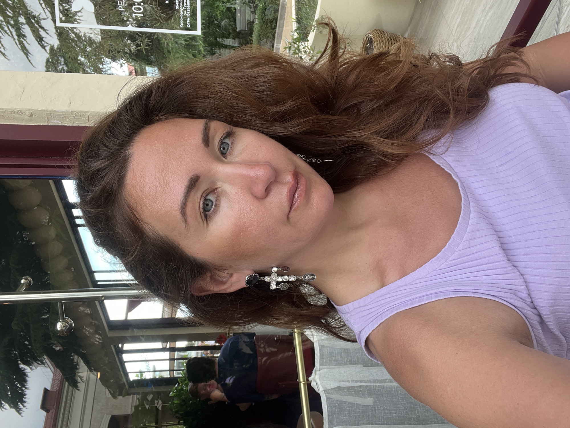 The image is a selfie taken by a woman with long, wavy brown hair and blue eyes. She is wearing a light purple ribbed tank top and a cross-shaped earring with charms. She is sitting at a table outdoors, with a window behind her that reflects trees and buildings. A sign in the window appears to be in Russian and has the number "10:00" on it. The woman is smiling slightly and looking at the camera. The background appears to be a cafe or restaurant with people seated at tables. The overall scene suggests a casual, daytime setting in an urban environment.