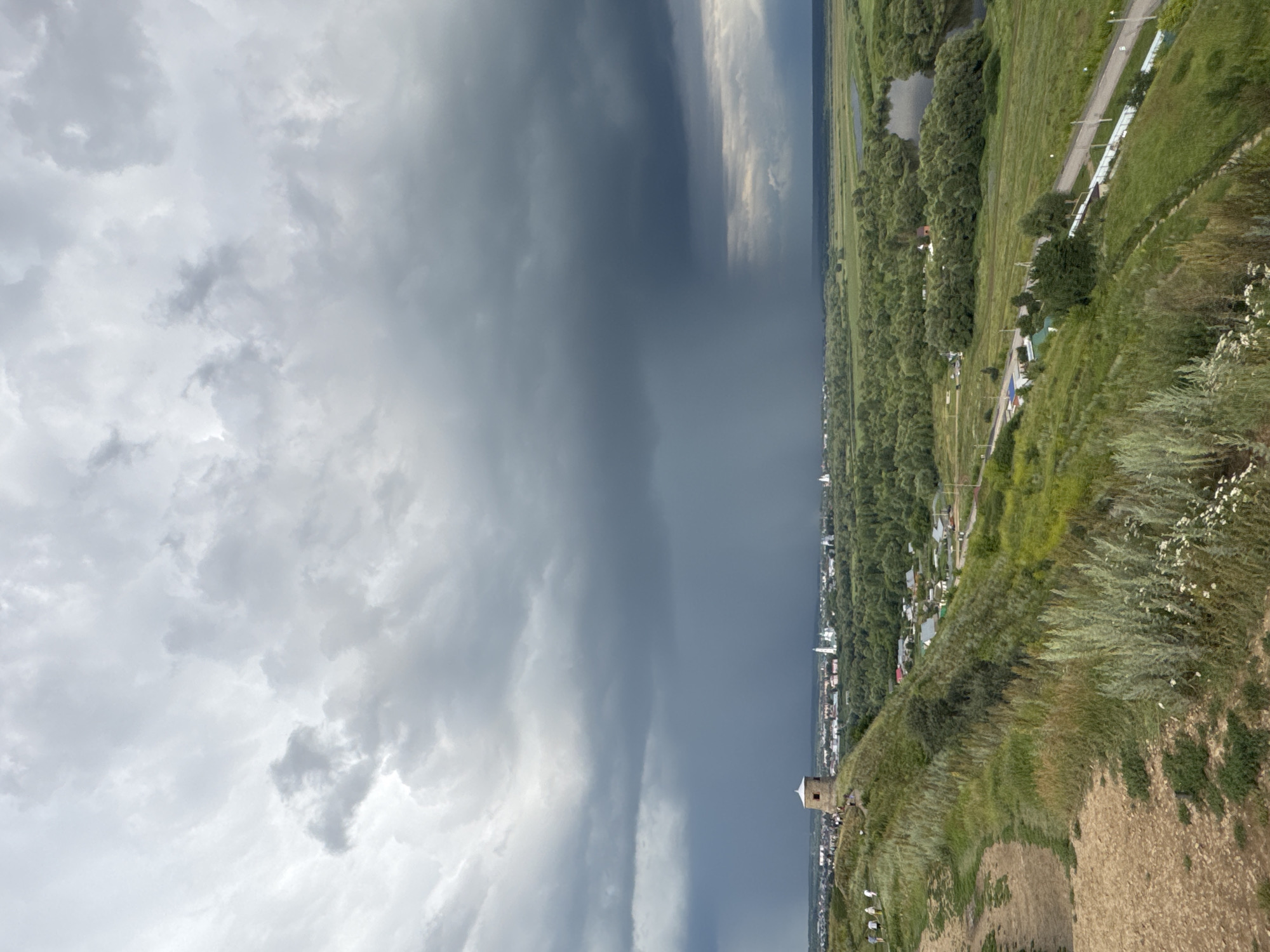 An aerial view captures the city of Yelabuga, Russia, under a dramatic, overcast sky. In the foreground, a grassy hill slopes down towards a valley. A path winds its way up the hill, leading to a circular stone structure with a conical roof, reminiscent of a tower or old mill. Several individuals are visible walking on the hill.
The middle ground reveals a cityscape with numerous buildings, including white structures with green roofs, likely churches, and smaller residential houses. Dense green foliage, representing trees and parks, blankets the area between the city and a winding river. A dark, heavy rain shower is actively falling in the distance, obscuring the horizon. The sky is filled with dark, ominous storm clouds.