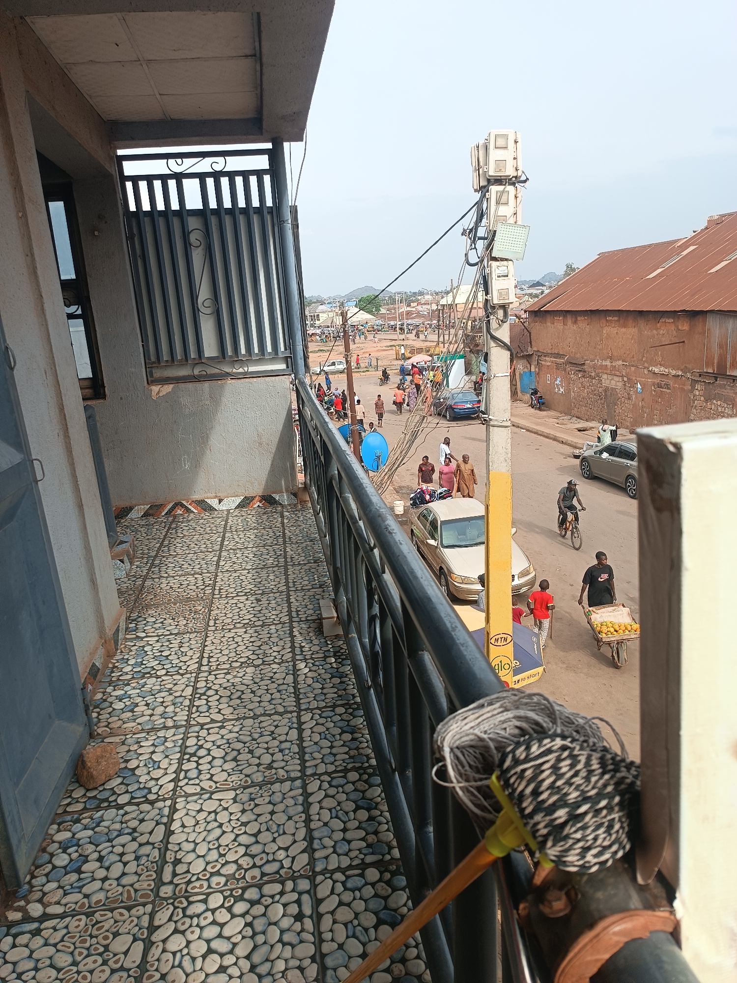 The image, taken from a balcony in Bukuru, Nigeria, shows a street scene. The balcony has a pebble-patterned floor, a metal railing, and a mop leaning against it. Below, the street is populated with people walking, riding bicycles, and pushing carts. There are several cars parked or moving on the road. A utility pole with electrical equipment is visible. Buildings with corrugated metal roofs and faded brick walls line the street. In the distance, there are more buildings and a hazy sky. The day appears overcast and the general environment suggests a residential and commercial area.