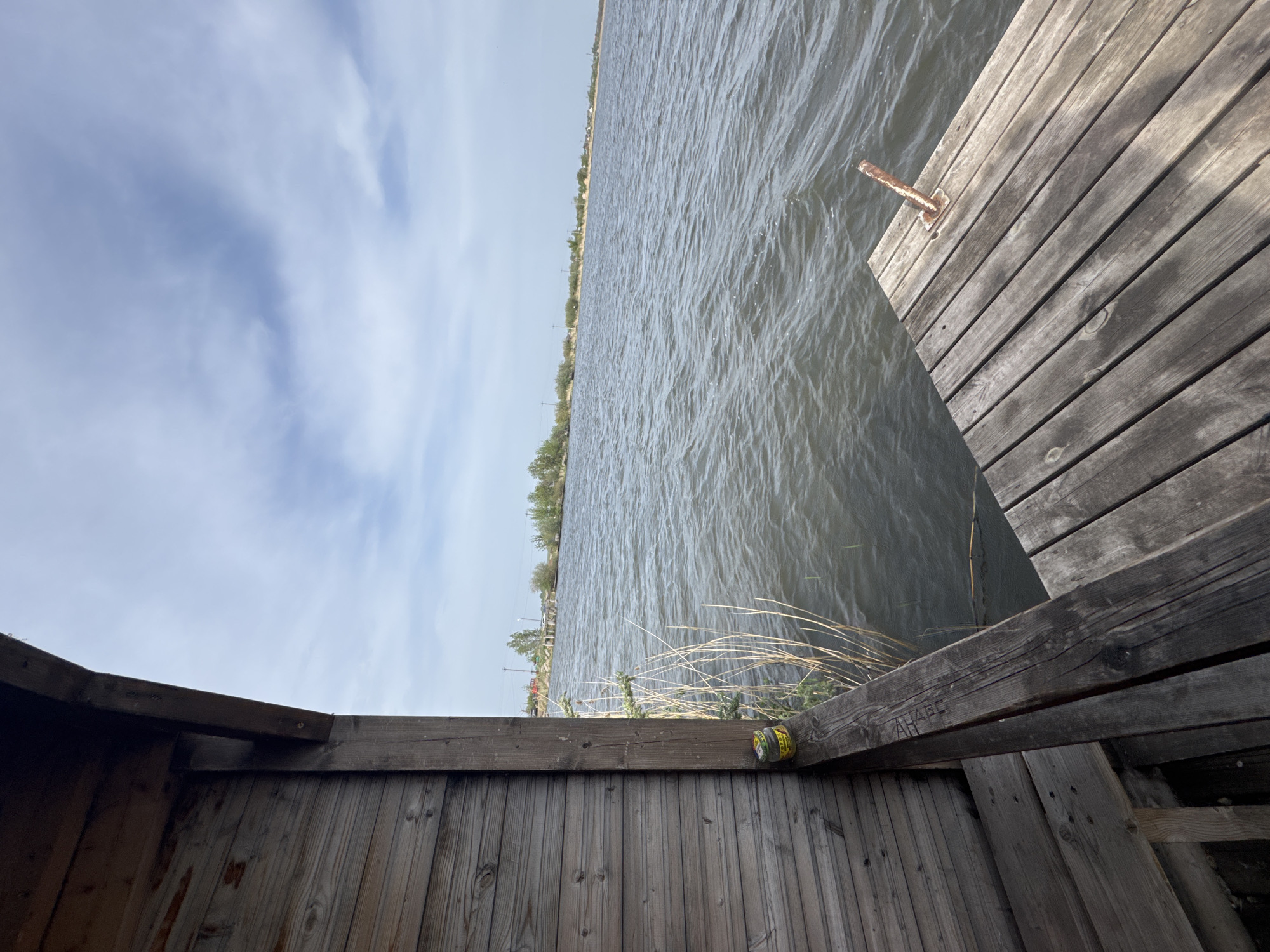 The image shows a view from a wooden structure, possibly a shed or small building, overlooking a body of water. The water appears to be a lake or wide river, with trees and possibly buildings visible on the opposite shore. A wooden dock extends into the water on the right side of the frame. The sky is partly cloudy. In the foreground, the wooden structure is visible, with a can resting on a beam. The overall impression is of a waterfront scene, possibly in a rural or suburban setting. No city or individuals are explicitly identifiable within the scene.
