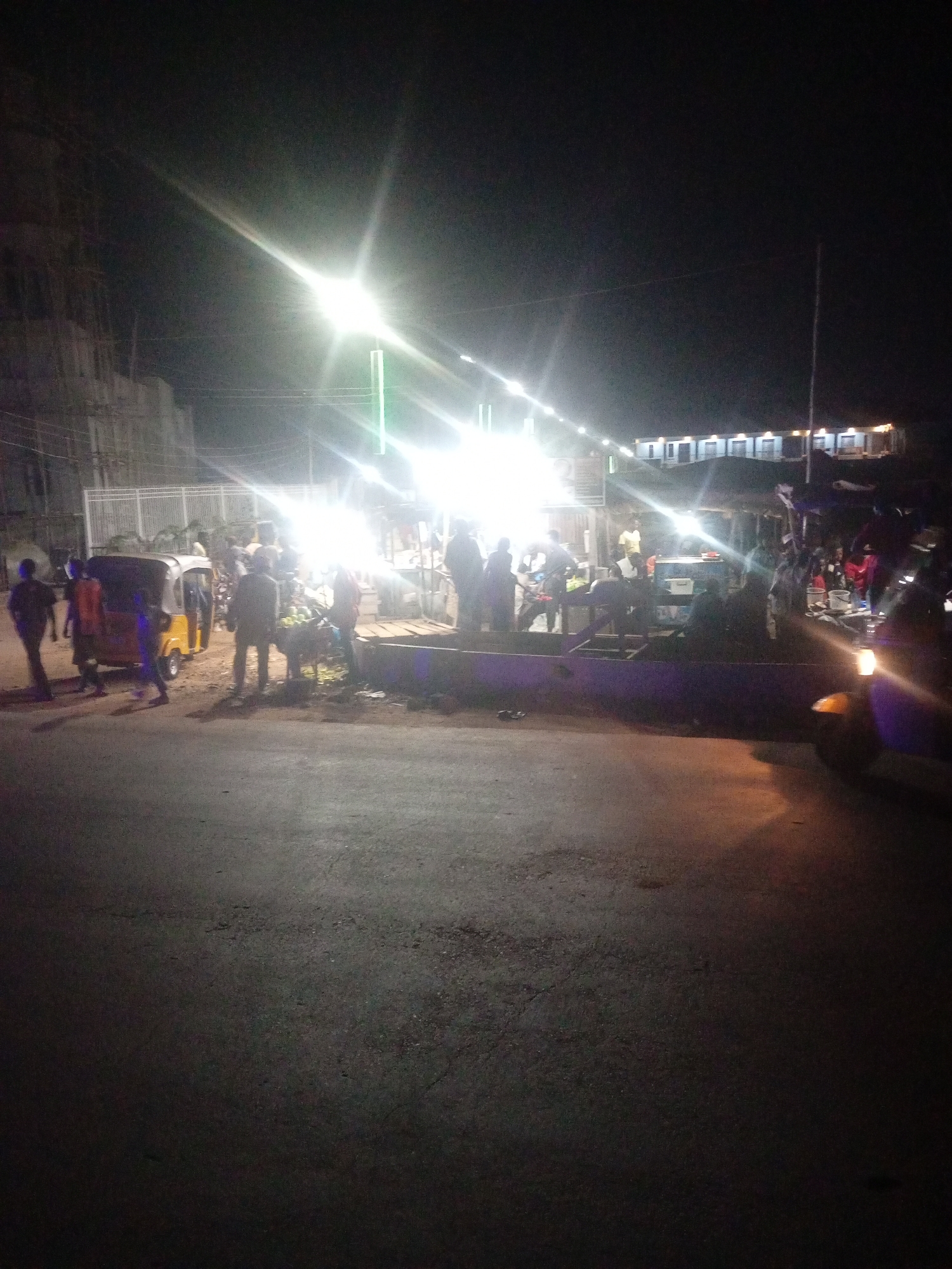The image captures a nighttime scene on a street in Tundun Wada West, Nigeria. The street is paved and appears to be an area with some commercial activity, possibly a market or roadside stalls. Bright overhead lights, likely streetlights or lights from businesses, illuminate the scene, creating strong glare and casting long shadows.
Several people are visible, some standing, some walking, and some gathered around what appear to be stalls. There are vehicles present, including a yellow auto-rickshaw (a tuk-tuk) parked on the side of the road and another vehicle partially visible on the right side of the frame. In the background, there are buildings, some of which appear to be under construction, indicated by scaffolding and unfinished elements. Another set of buildings in the distance appears to be shops or residences with illuminated windows. The overall atmosphere suggests a bustling but dimly lit urban environment at night.