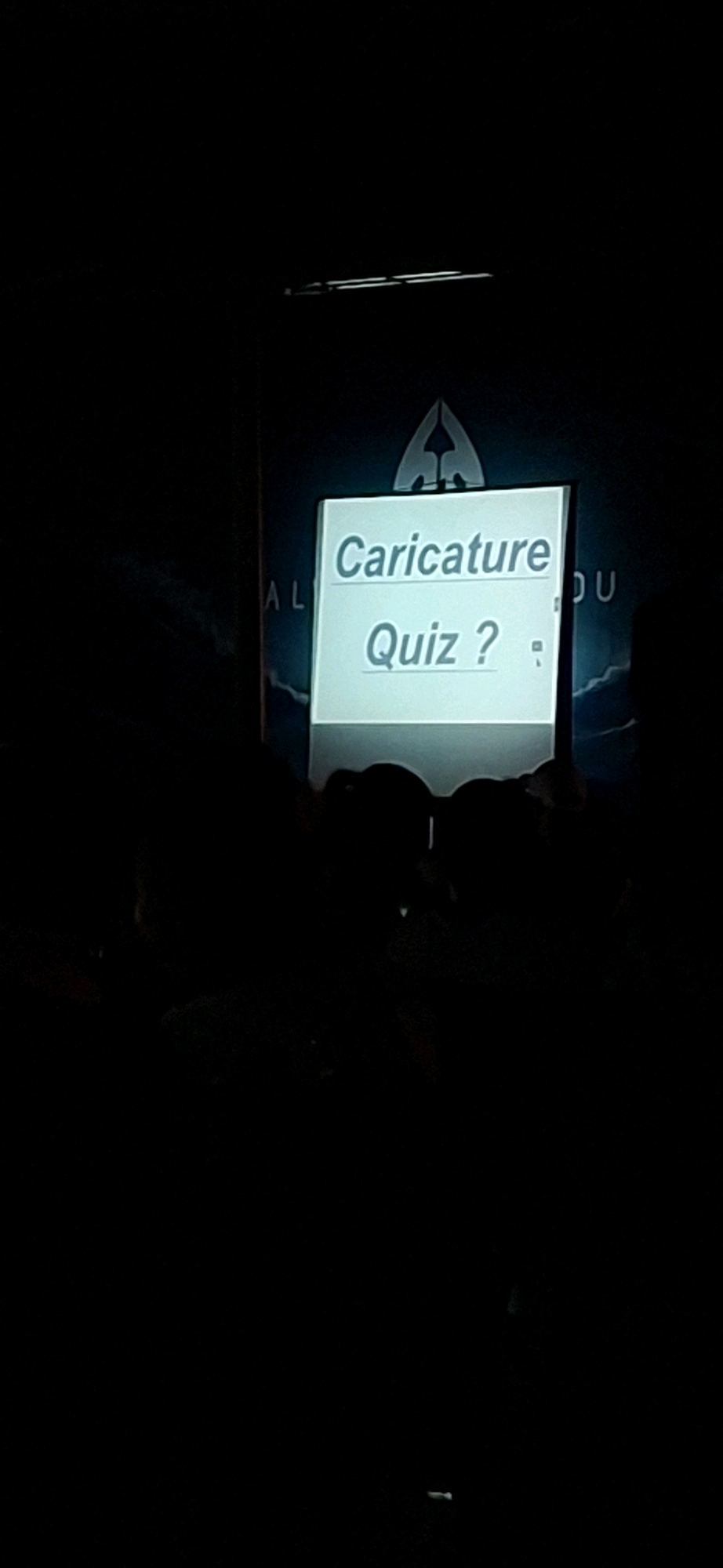 The image shows a projection screen with the words "Caricature Quiz?" displayed in a light blue font. The screen is framed by a darker material with a subtle design at the top. Partially visible on the left side of the screen are the letters "AL" in a dark blue font. To the right of the screen, the letters "DU" are also visible in the same dark blue font. The overall lighting suggests a dimly lit indoor environment, possibly a presentation or event. The location in Kemer, Turkey is not discernible from the visual information in the image.