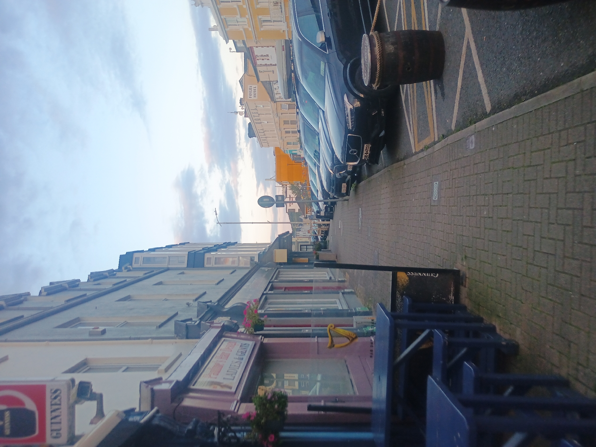 The image depicts a street scene in a town or city, likely during overcast weather given the soft lighting. On the left, a light blue building with a prominent Guinness sign is visible. Below the sign, a shop window with the words "Pauline's Hair Design LADIES & GENTS" is partially obscured. Blue outdoor tables and seating are in the foreground. The sidewalk is paved with rectangular bricks, and a row of parked cars lines the right side of the street. The cars are of various dark colors, including black SUVs. Further down the street, several buildings with multiple stories are visible, one of which is yellow and has the sign "FOYLES HOTEL". A street lamp is present on the left side of the image. A wooden barrel is positioned on the street, near the parked cars. The sky is cloudy, with hints of pink and orange, suggesting either dawn or dusk.