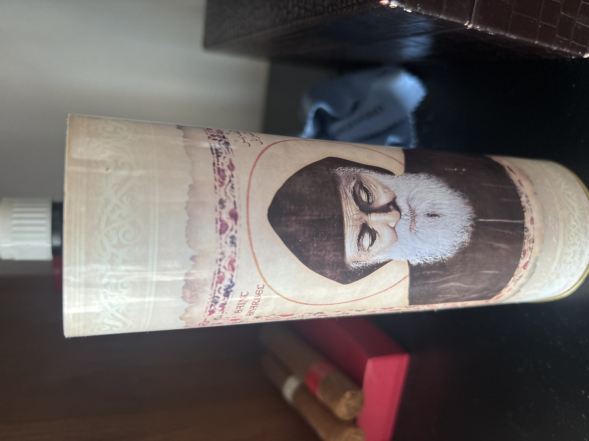 A tall, cylindrical candle with an image of Saint Charbel is the central focus of the image. The candle has an ornate, cream-colored paper wrapper adorned with intricate patterns and Arabic calligraphy. The image on the candle depicts a saintly figure with a beard and a monastic hood. To the left of the candle, two cigars are visible, resting in a red box. The background is blurred, but suggests an indoor setting. The candle is placed on a dark, reflective surface, likely a table. The image is taken from a slightly elevated angle, looking down at the candle. There is no indication that this image was taken in Dubai, United Arab Emirates, as the scene depicted does not contain any recognizable landmarks or context clues to suggest this location.