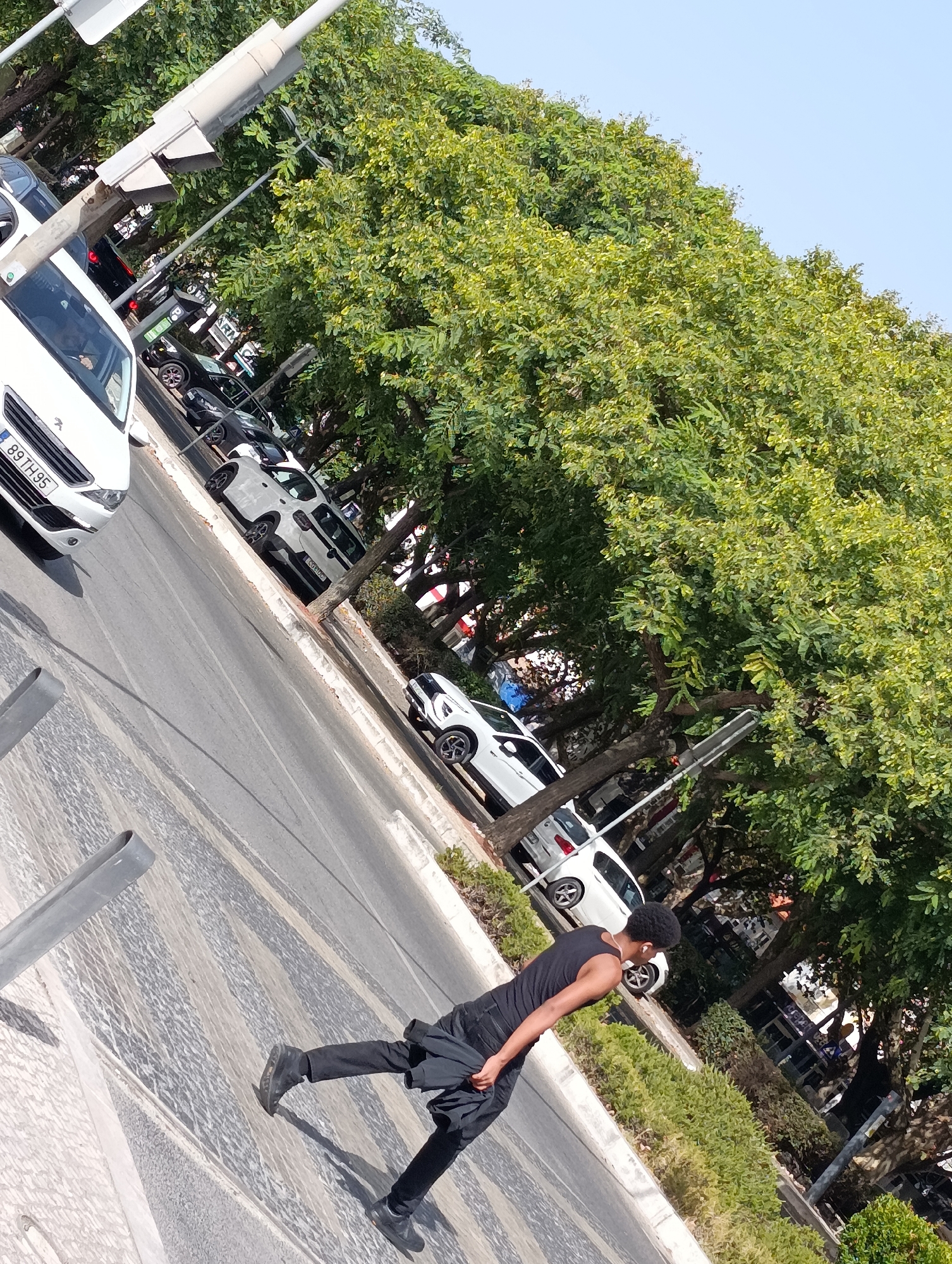 The image depicts a street scene in Setúbal, Portugal. A young Black man in a black tank top and black pants is walking across a pedestrian crossing. He is looking towards his right. The street is lined with trees and parked cars. A white car with the license plate "89 TH95" is in the foreground on the left. A traffic light pole is visible in the upper left of the image. The sky is clear and blue.