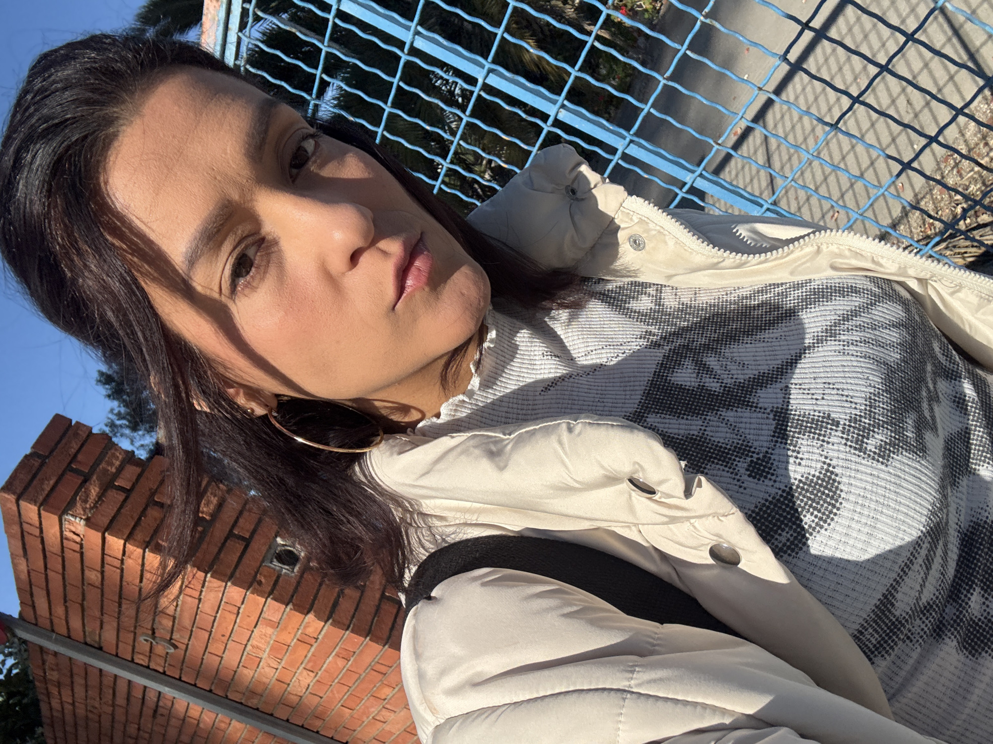 A woman with dark hair and gold hoop earrings is shown in a close-up, tilted selfie taken outdoors in Barcelona, Spain. She wears a beige puffer jacket over a gray and white patterned top, and a black backpack strap is visible. Behind her is a blue metal fence with shadows cast on the ground. A portion of a brick wall is also visible. The lighting suggests it is daytime.