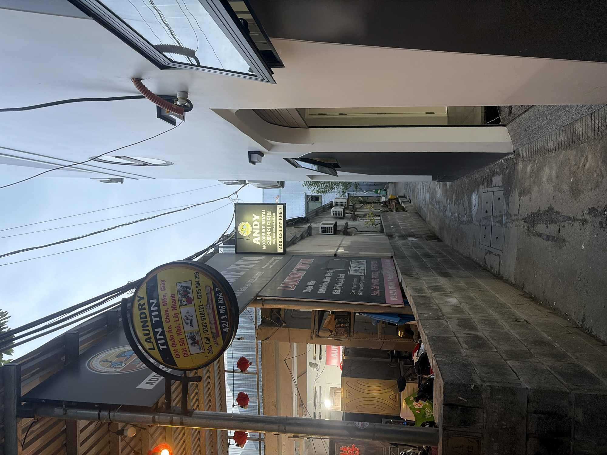 The image depicts a narrow alleyway in the city of Đà Nẵng, Vietnam, likely at dusk or in the early evening, indicated by the artificial lighting and the dim sky. On the left side of the alley, a laundry business named "LAUNDRY TIN TIN" is prominently displayed with a circular sign featuring Vietnamese text and images of washing machines and clothing. Next to it, a vertical sign for "ANDY MOTORBIKE RENTAL" is visible, also with Vietnamese and Korean text. The laundry establishment appears to have washing machines stacked inside, suggesting an operational business. The alleyway itself is paved with concrete, and a low concrete wall borders the left side. Various electrical wires are strung overhead. On the right side of the alley, a modern building with white walls and black-trimmed windows dominates the scene. An arched doorway is visible on this building. The overall impression is a utilitarian urban environment with commercial signage typical of a local neighborhood in Vietnam.