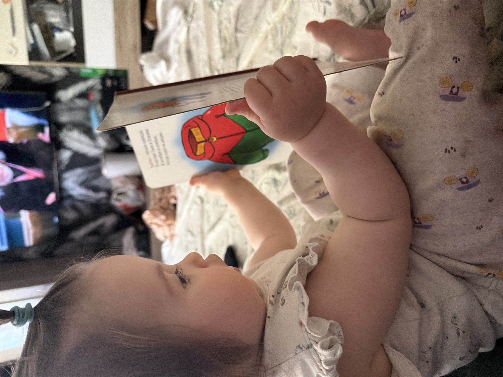 A young child is lying on a bed, holding a book open. The child is facing toward the left of the frame and holding the book above their head. The book has a cartoon drawing of a red and green object, potentially clothing, on the open page. The child is wearing a light-colored outfit with a ruffle detail at the shoulder. The bed has a patterned sheet. Behind the bed is a TV and other items, some blurred, in a room. It's not possible to confirm the location as Staraya, Russia, based on the given image.