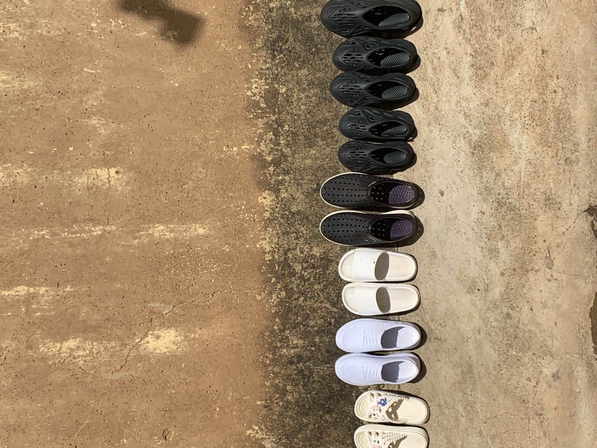 The image displays a forensic scene in Rafin Par, Nigeria, featuring a collection of 12 individual shoes, arranged as six pairs, positioned horizontally along the base of a textured wall on a ground surface.
The background consists of a vertical, reddish-brown earthen or rendered wall, exhibiting significant weathering with areas of lighter, yellowish-beige discoloration, cracks, and granular texture. A dark shadow is cast onto the upper right portion of the wall, originating from an unseen object or structure to the right.
The ground surface beneath the shoes is a lighter, sandy-brown or light grey granular material, possibly a mix of dirt and weathered concrete, showing varying textures and small darker patches.
From left to right, the shoes are as follows:
1. A pair of white, woven-textured slip-on sandals. The left sandal features a small blue flower emblem, and the right sandal has a pink flower emblem.
2. A pair of plain white, open-toed, slip-on slide sandals with flat soles.
3. A pair of white, closed-toe, lace-up style shoes, appearing to be made of a smooth, possibly synthetic material rather than traditional canvas.
4. A pair of black, closed-toe, slip-on clogs or water shoes, characterized by numerous circular perforations on the upper and sides, with visible white outer soles.
5. A pair of black, molded, futuristic-style clogs or slippers, featuring large, organically shaped cutouts on the upper surface.
6. An identical pair to the fifth, consisting of black, molded, futuristic-style clogs or slippers with organically shaped cutouts.
All shoes are uniformly oriented with the toes facing away from the viewer. The scene is illuminated by bright, natural light, casting distinct shadows beneath each pair of shoes on the ground surface. No people are visible in the frame.