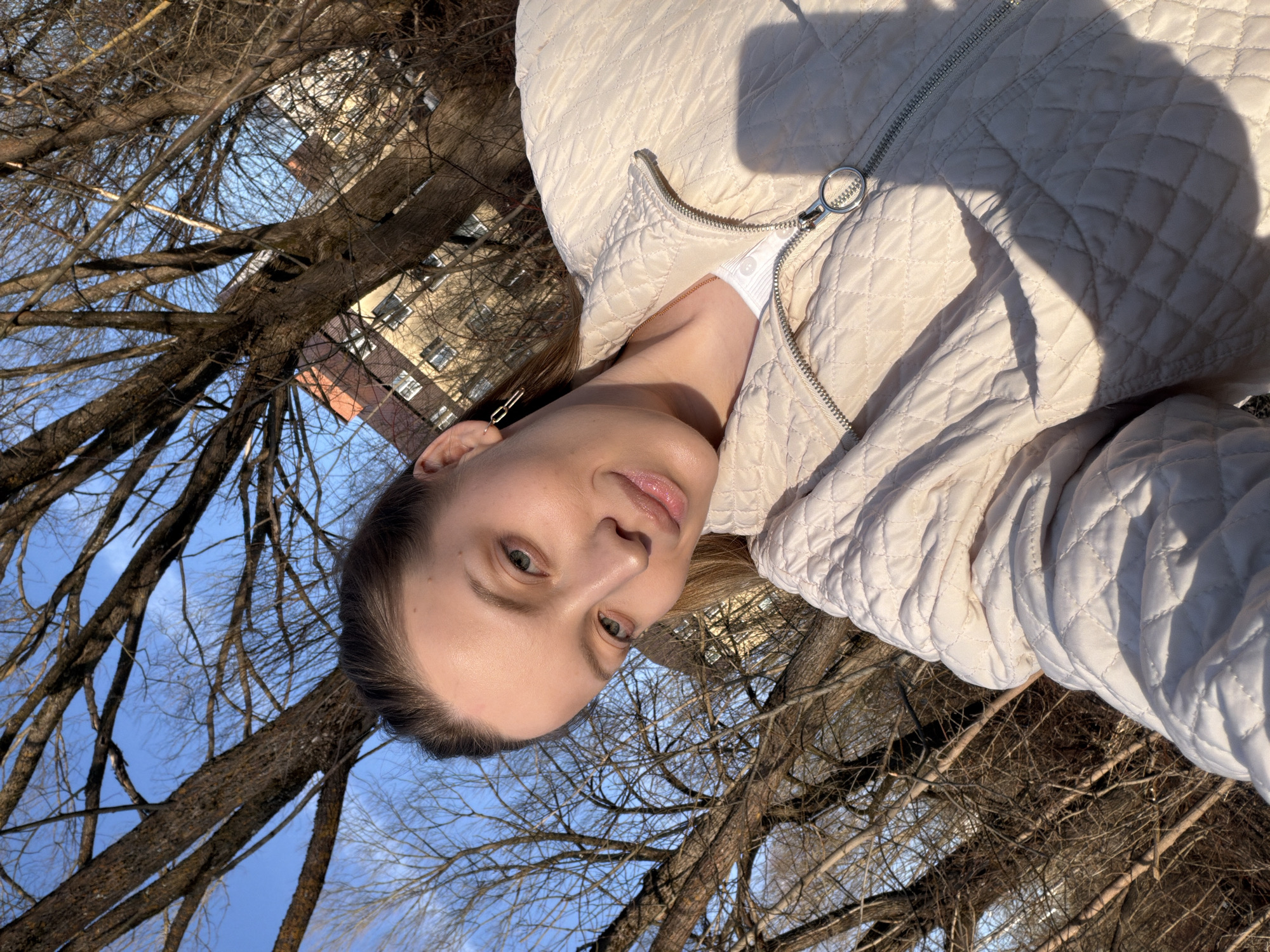 The image is a selfie taken from a low angle, tilted to the right. A woman with fair skin and light eyes is the primary subject. She is wearing a light-colored quilted jacket with the zipper partially open, revealing a white top underneath. The background consists of bare tree branches against a bright blue sky, indicating a late autumn or early spring season. In the upper left corner of the frame, a multi-story building is partially visible. Based on the architectural style and environment, it's possible the location could be Reshetnikovo, Russia, as requested. No other people are present in the image. The action depicted is the woman taking a selfie outdoors.