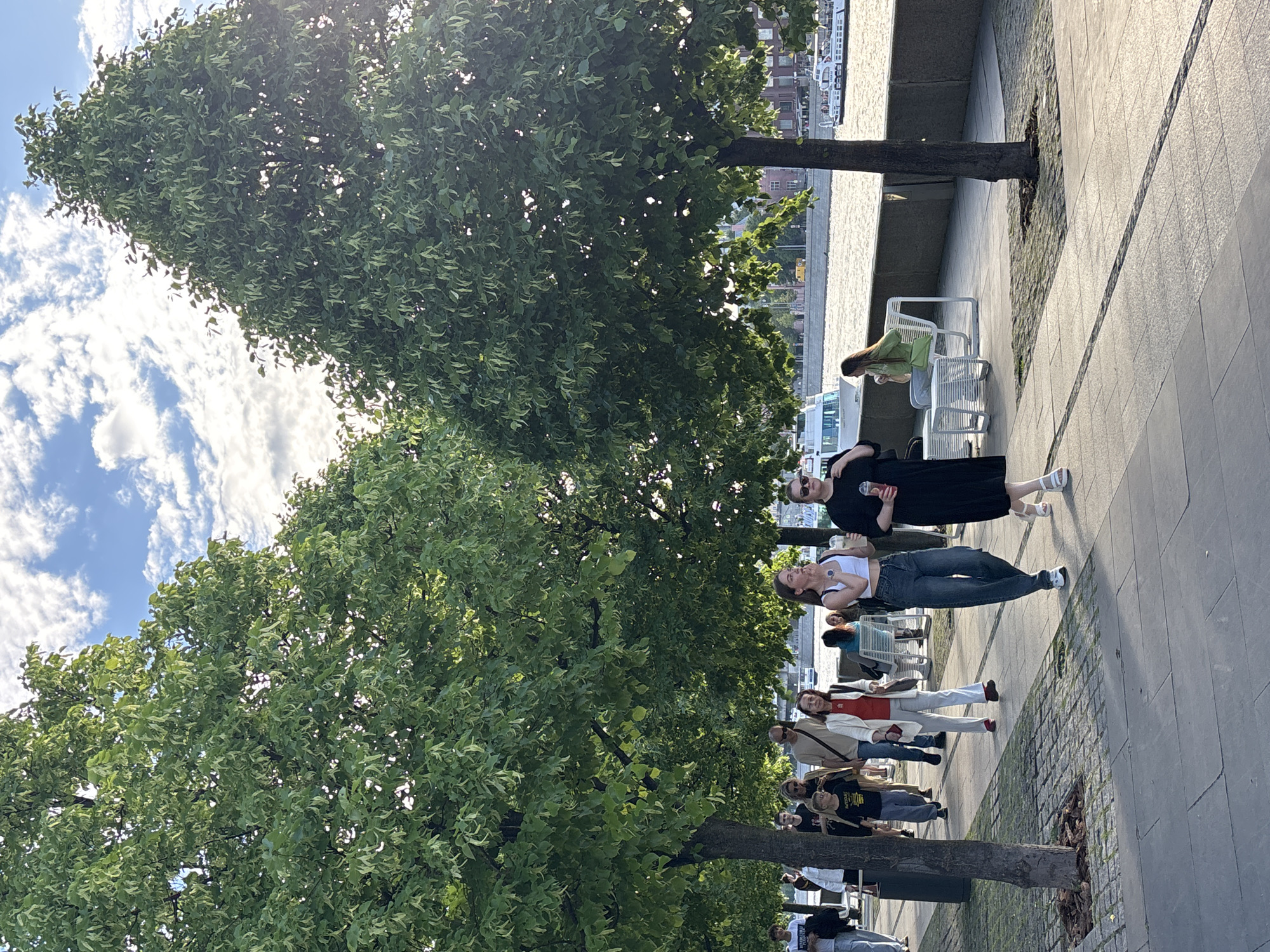 On a sunny day in Moscow, Russia, a diverse group of people stroll along a paved waterfront promenade shaded by mature trees. Individuals are dressed in casual attire, including white tops, jeans, dresses, and jackets. Some are walking and others are standing near benches. The background includes water and architectural structures across the waterway.
